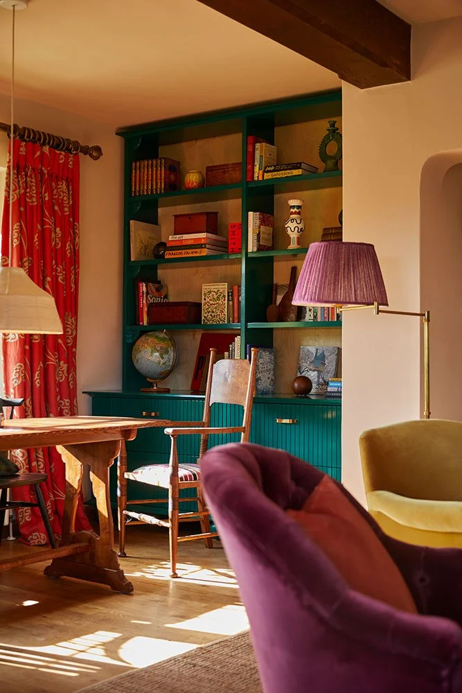 Eclectic living room designed by Coal Cornwall, featuring an emerald green built-in bookcase with lower cabinetry, vibrant red patterned curtains, a purple plush armchair, a brass floor lamp with a pleated shade, and a vintage wooden desk chair.