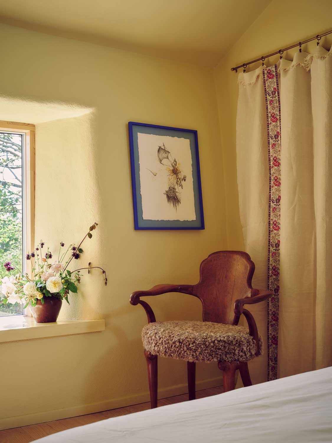 Warm bedroom corner designed by Coal Cornwall featuring pale yellow plaster walls, an antique wooden armchair with textured shearling upholstery, floral-trimmed curtains, and a blue-framed artwork.