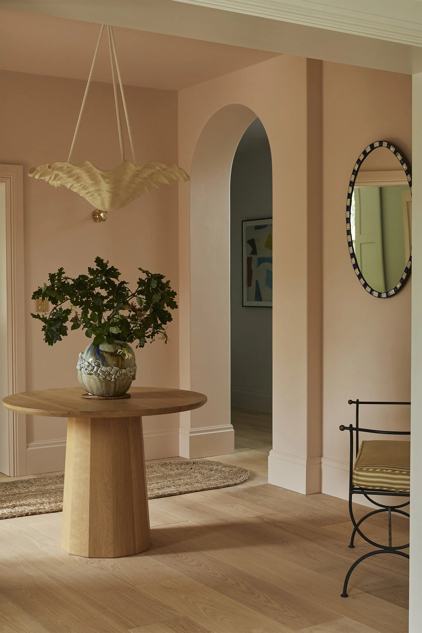 Elegant entryway designed by Coal Cornwall, featuring soft terracotta walls, an arched doorway, a central round oak pedestal table with a leafy vase, a sculptural rope-pendant light, and light oak floorboards.