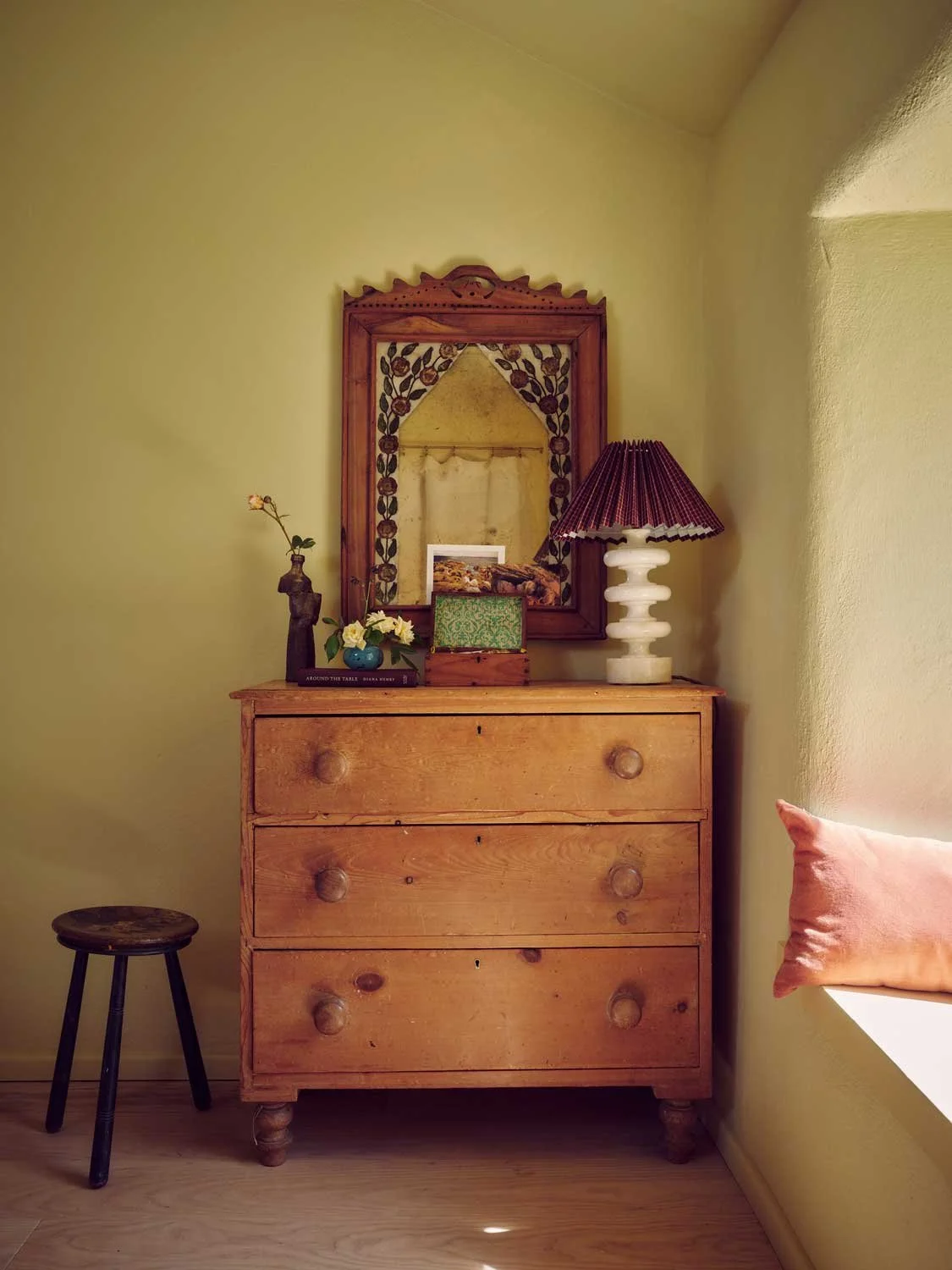Cozy bedroom corner designed by Coal Cornwall, featuring an antique pine chest of drawers, an ornate hand-carved wooden mirror, a stacked ceramic lamp base with a pleated shade, and a dark wooden tripod stool.