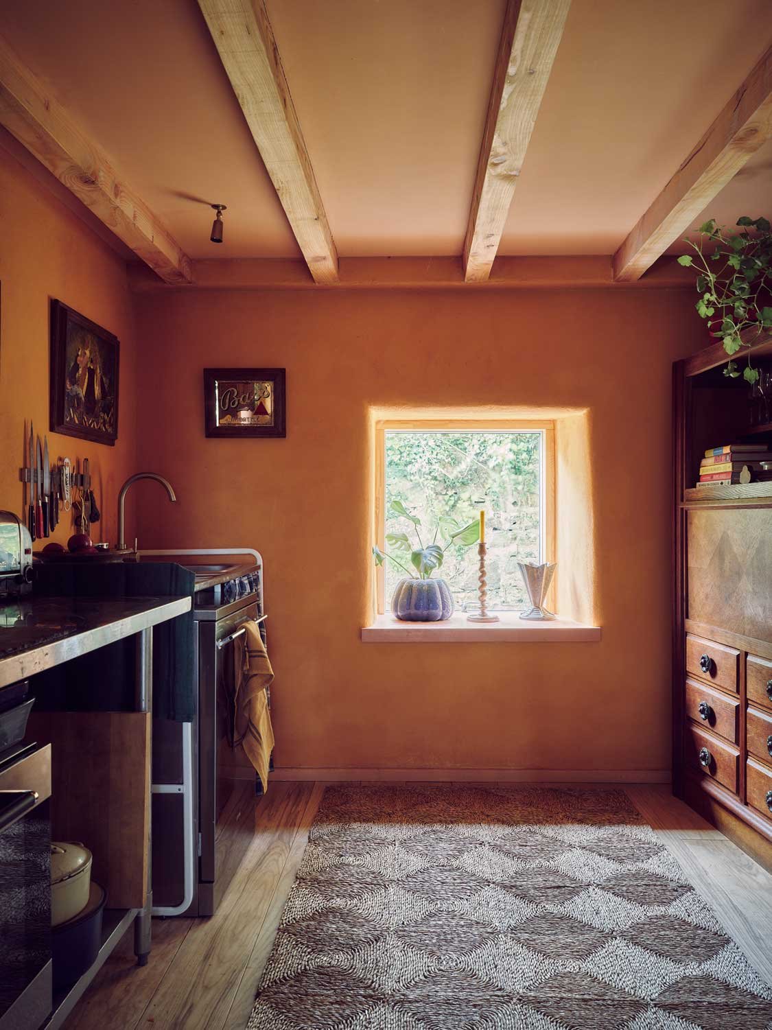 Rustic Cornish kitchen interior by Coal Cornwall featuring exposed timber ceiling beams, terracotta plaster walls, deep window sill with greenery, geometric runner rug, and freestanding wooden cabinetry.
