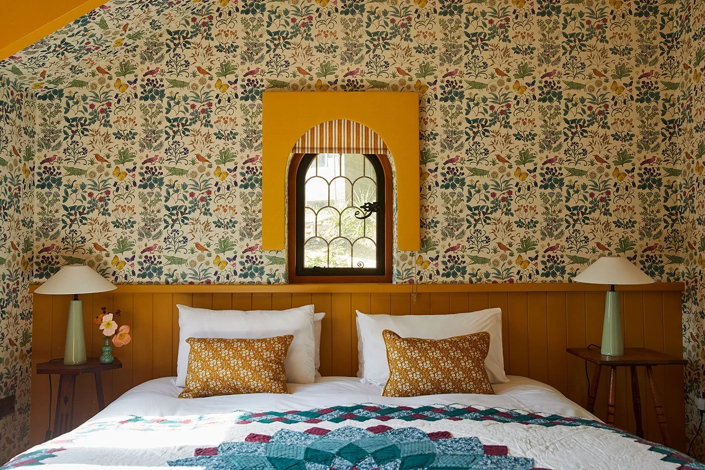 Whimsical bedroom designed by Coal Cornwall, featuring botanical insect-patterned wallpaper, a mustard yellow tongue-and-groove headboard, a central arched window casing, and sage green bedside lamps.