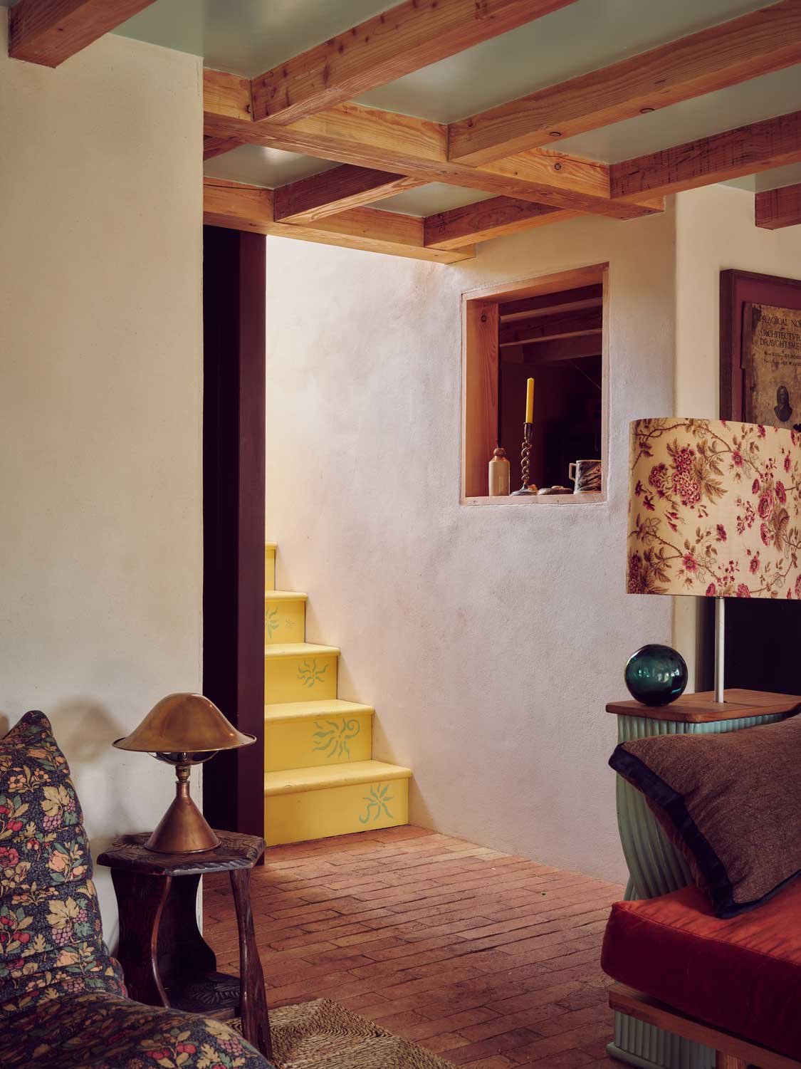 Cornish interior architecture by Coal Cornwall featuring hand-painted yellow stairs with floral motifs, exposed wooden ceiling beams, terracotta brick flooring, a floral lampshade, and a internal window opening.