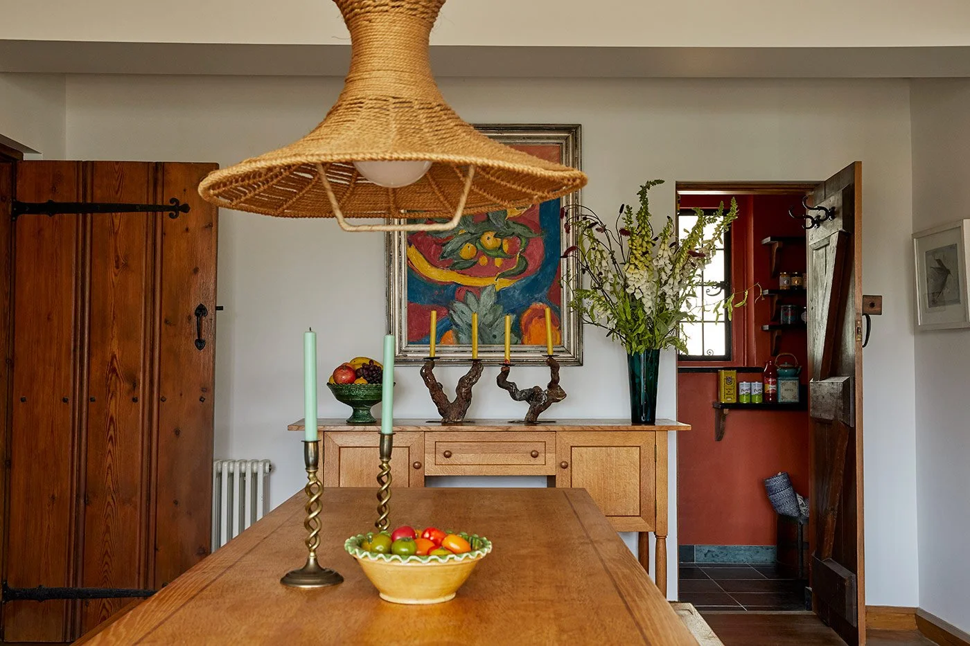 Rustic dining room designed by Coal Cornwall, featuring a wooden dining table with barley-twist brass candlesticks, a woven jute pendant light, a pine sideboard, colorful oil painting, and an open doorway leading to a terracotta-red pantry.