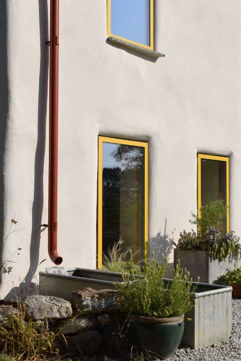 Cornish architectural exterior design by Coal Cornwall, featuring bright yellow window frames set against a smooth white rendered facade, paired with a terracotta downpipe, stone wall, and galvanised trough planters.