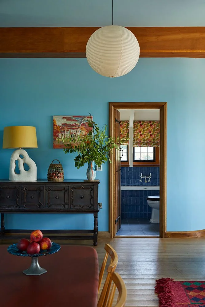 Vibrant sky blue living room designed by Coal Cornwall, featuring a dark carved sideboard, sculptural ceramic lamp, paper sphere pendant, and an open doorway revealing a cobalt blue tiled bathroom.