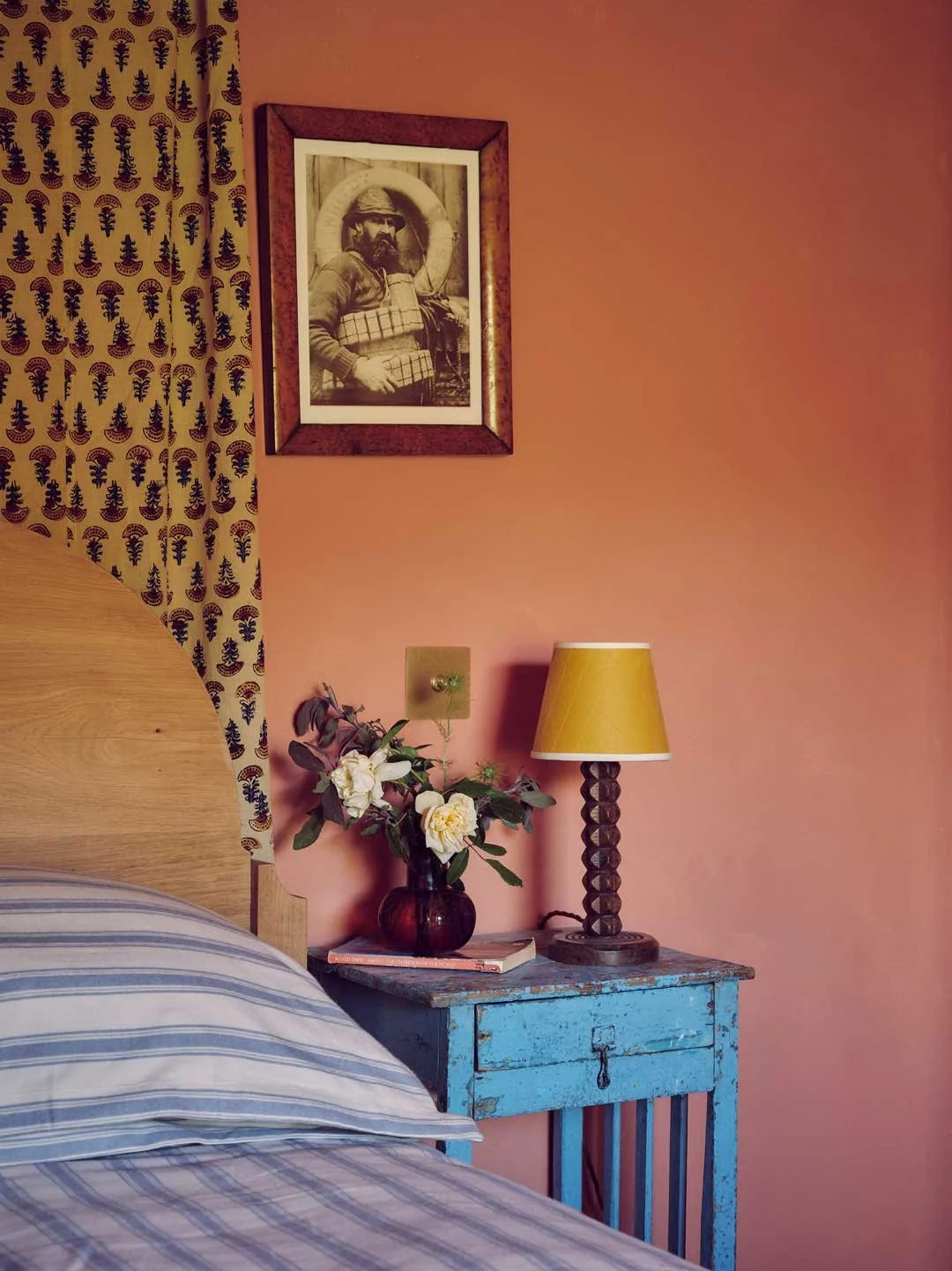 Cornish interior design by Coal Cornwall featuring a cozy bedroom corner with a natural wood headboard, striped bedding, vintage distressed blue nightstand, yellow lamp, and black-and-white art set against a dusty pink wall.