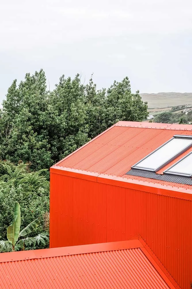 Elevated architectural view by Coal Cornwall showing an orange corrugated metal pitched roof equipped with dual Velux skylights, set against dense green foliage and rolling countryside views.