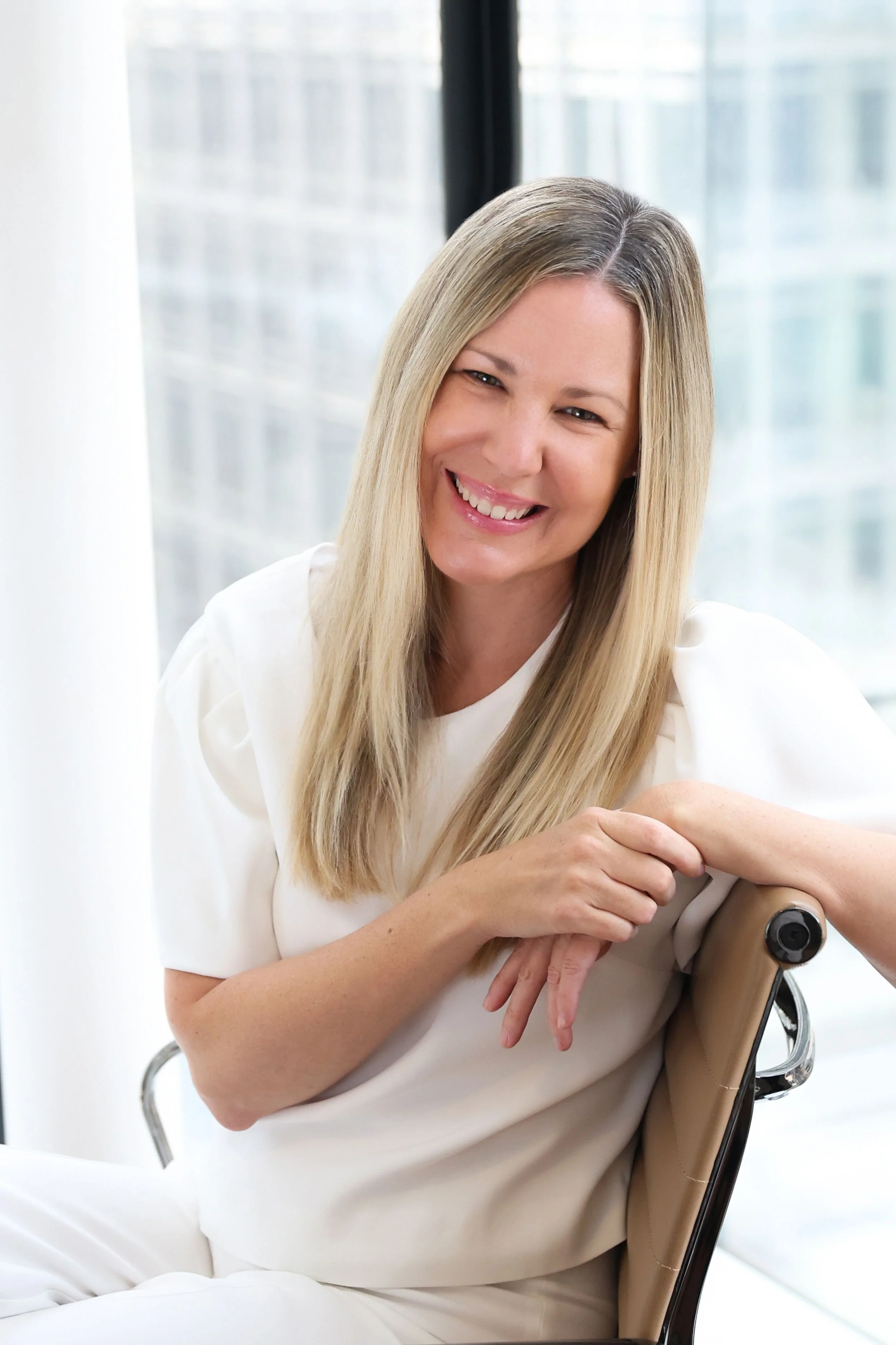 A woman with long blonde hair, smiling and sitting on a chair, wearing a white outfit with a modern background with large windows.