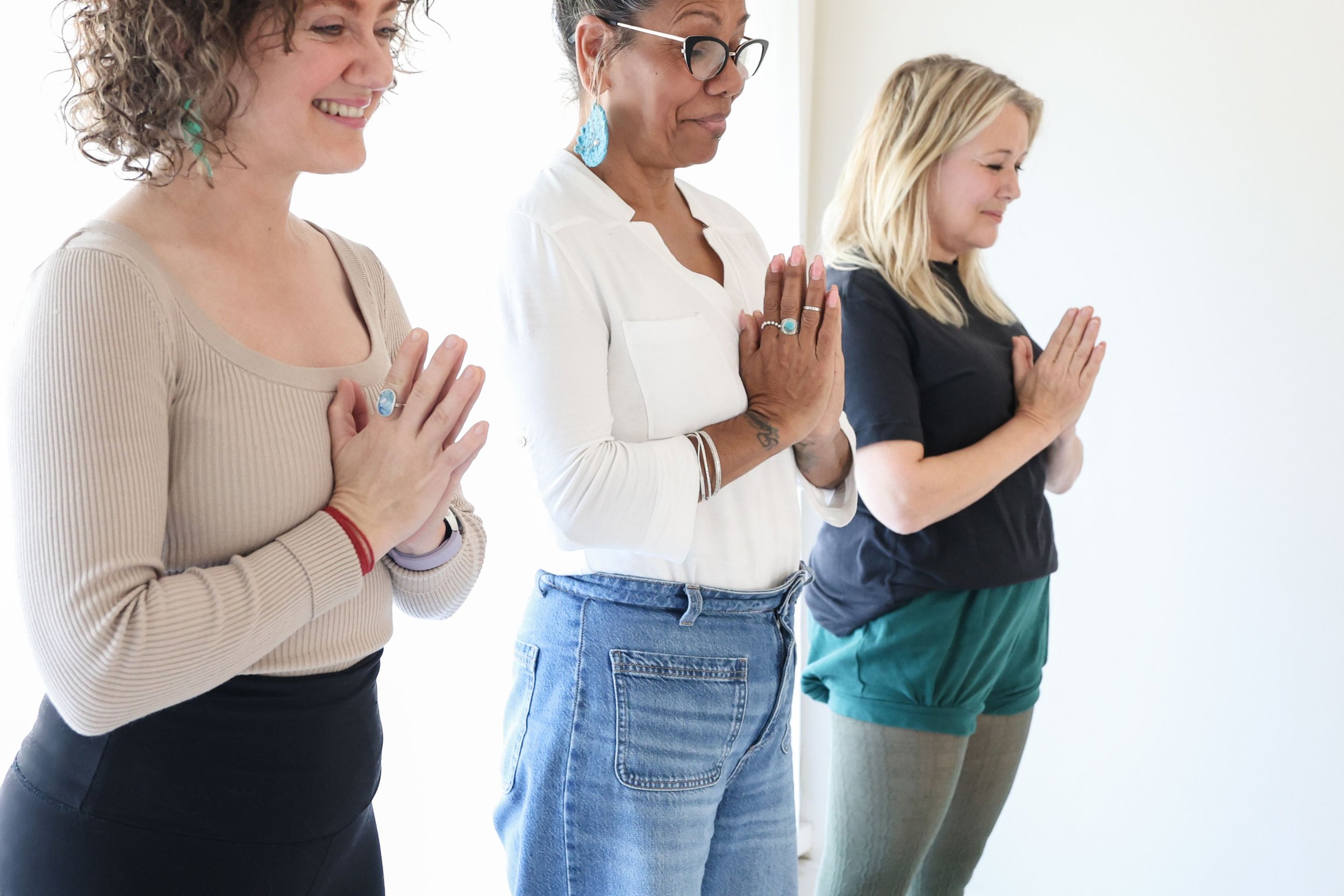 Three women standing in a row with hands in a prayer position, meditating or practicing yoga indoors.