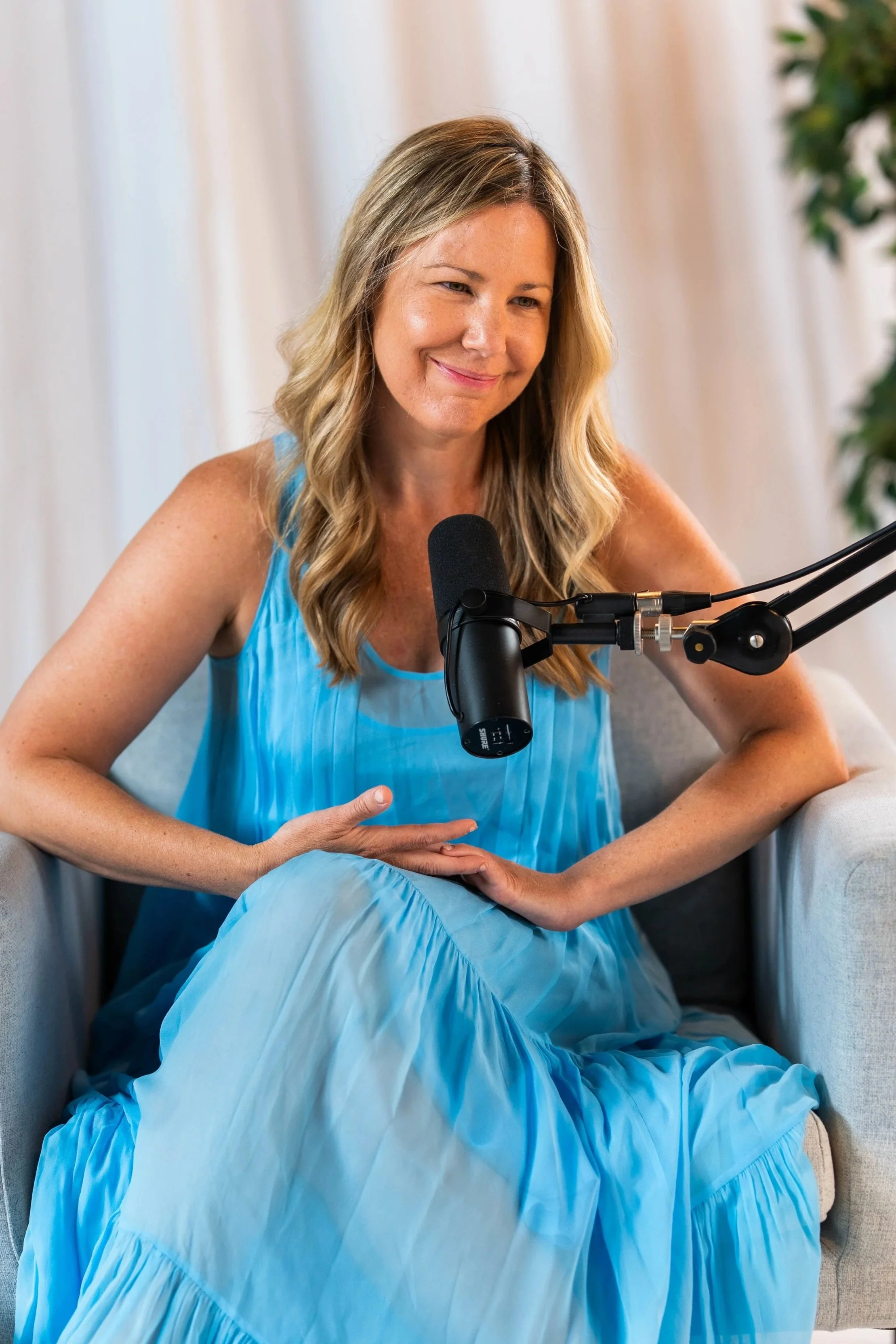 Woman in a blue dress sitting on a couch, smiling, with a microphone positioned near her face.