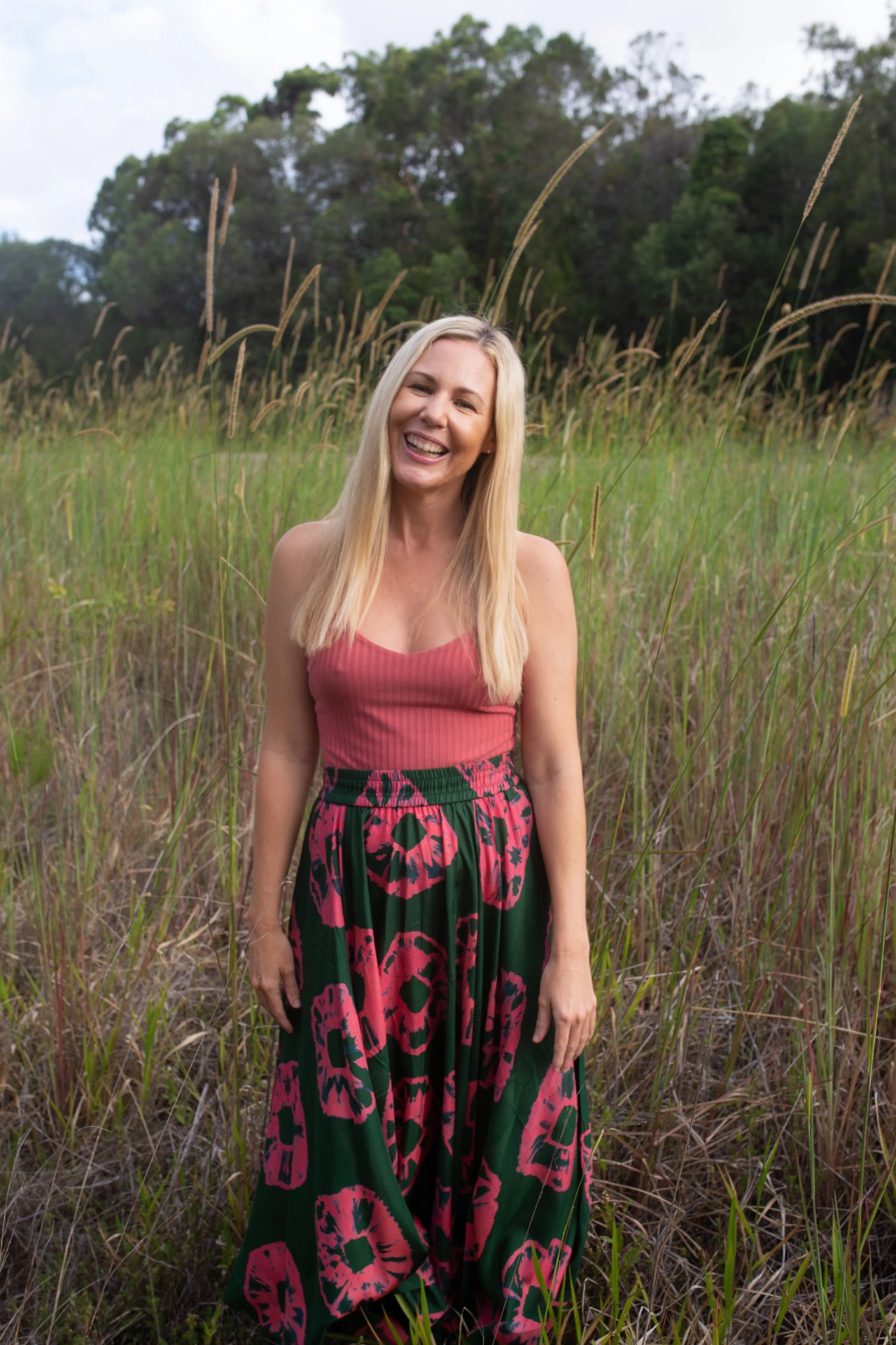 A smiling woman in a pink tank top and a long green skirt with pink flower patterns, standing in a grassy field with trees in the background.