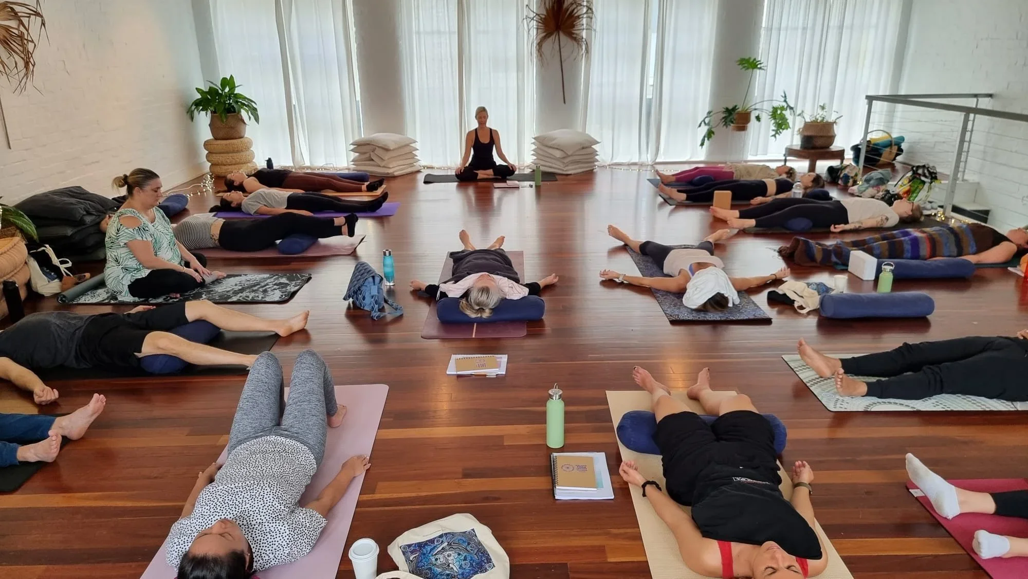 People participating in a yoga or meditation class lying on yoga mats with a teacher sitting cross-legged at the front of the room.