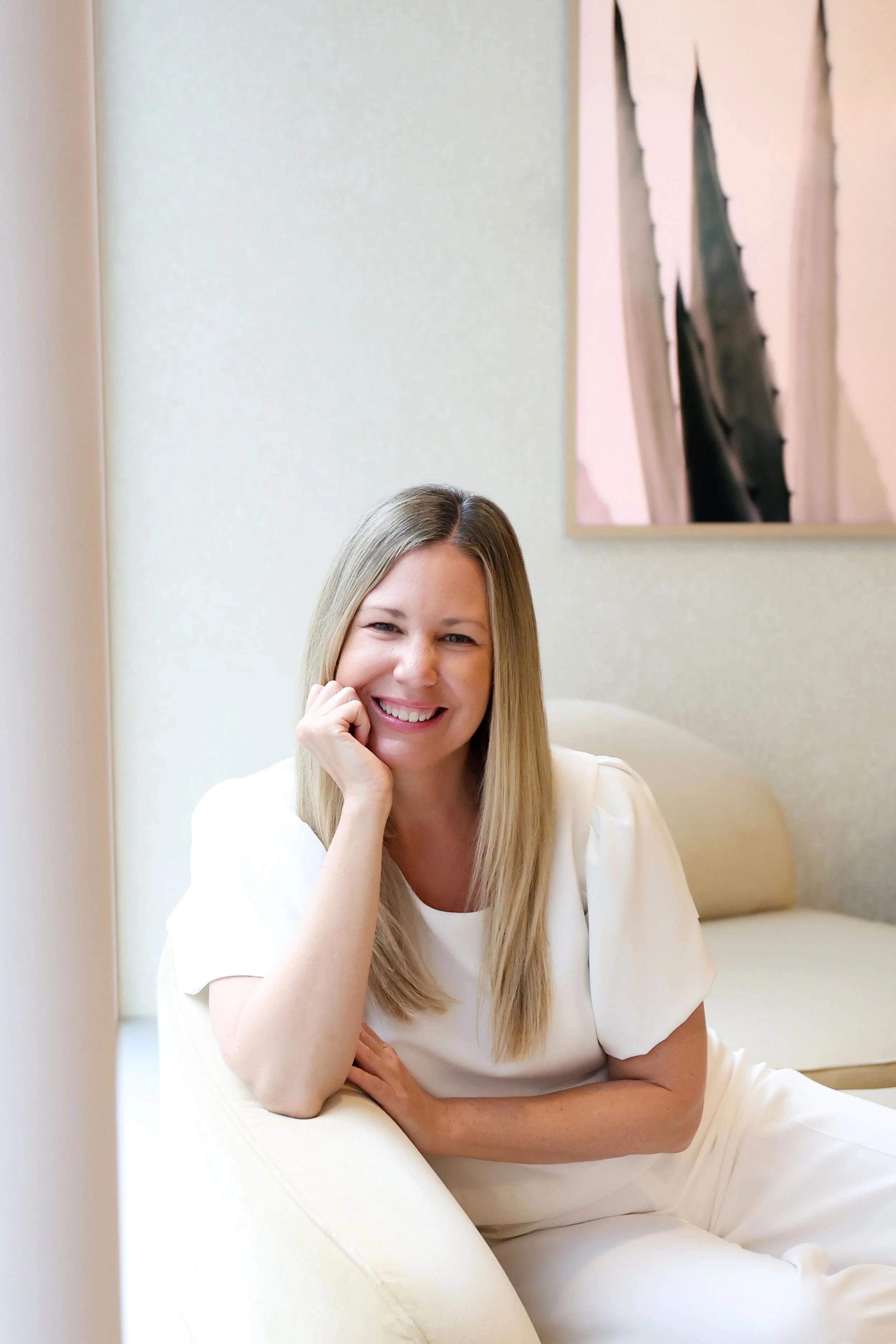 A woman with long blond hair smiling while sitting on a cream-colored sofa in a room with light-colored walls and abstract artwork in the background.