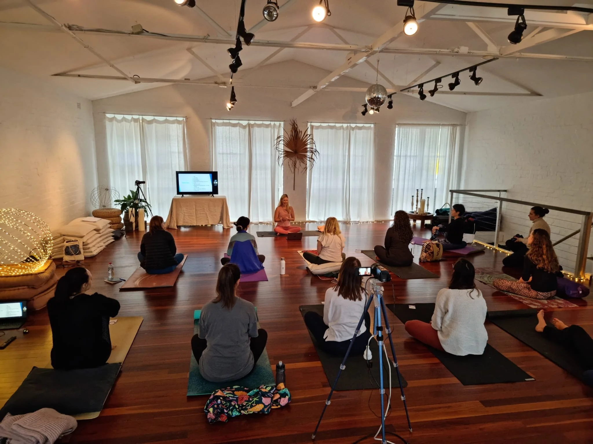 Group of people participating in a yoga or meditation class in a bright room with hardwood floors, white brick walls, and large window drapes.