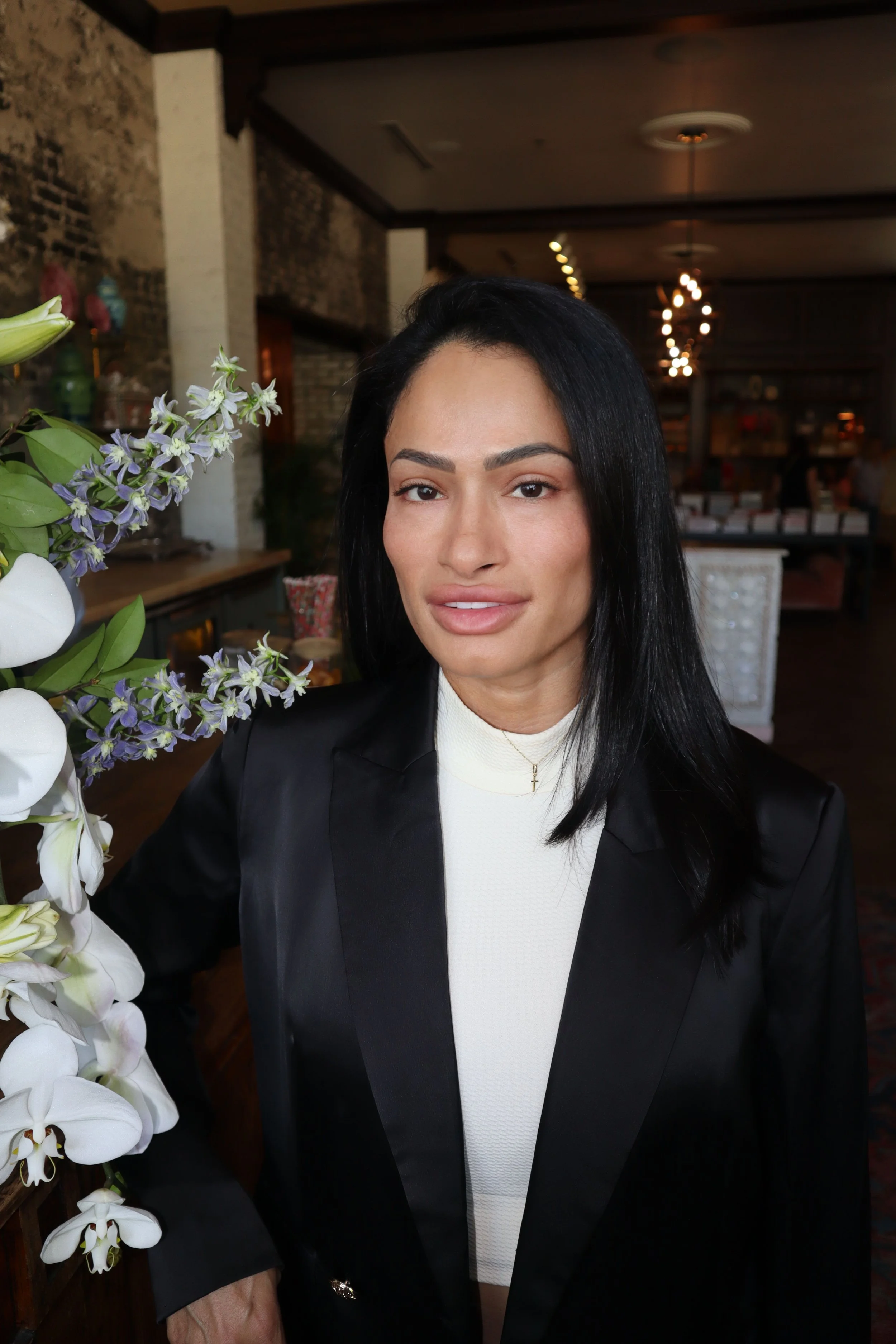 A woman with black hair and tan skin standing indoors, wearing a white turtleneck and black blazer, near a flower arrangement with white and purple flowers.