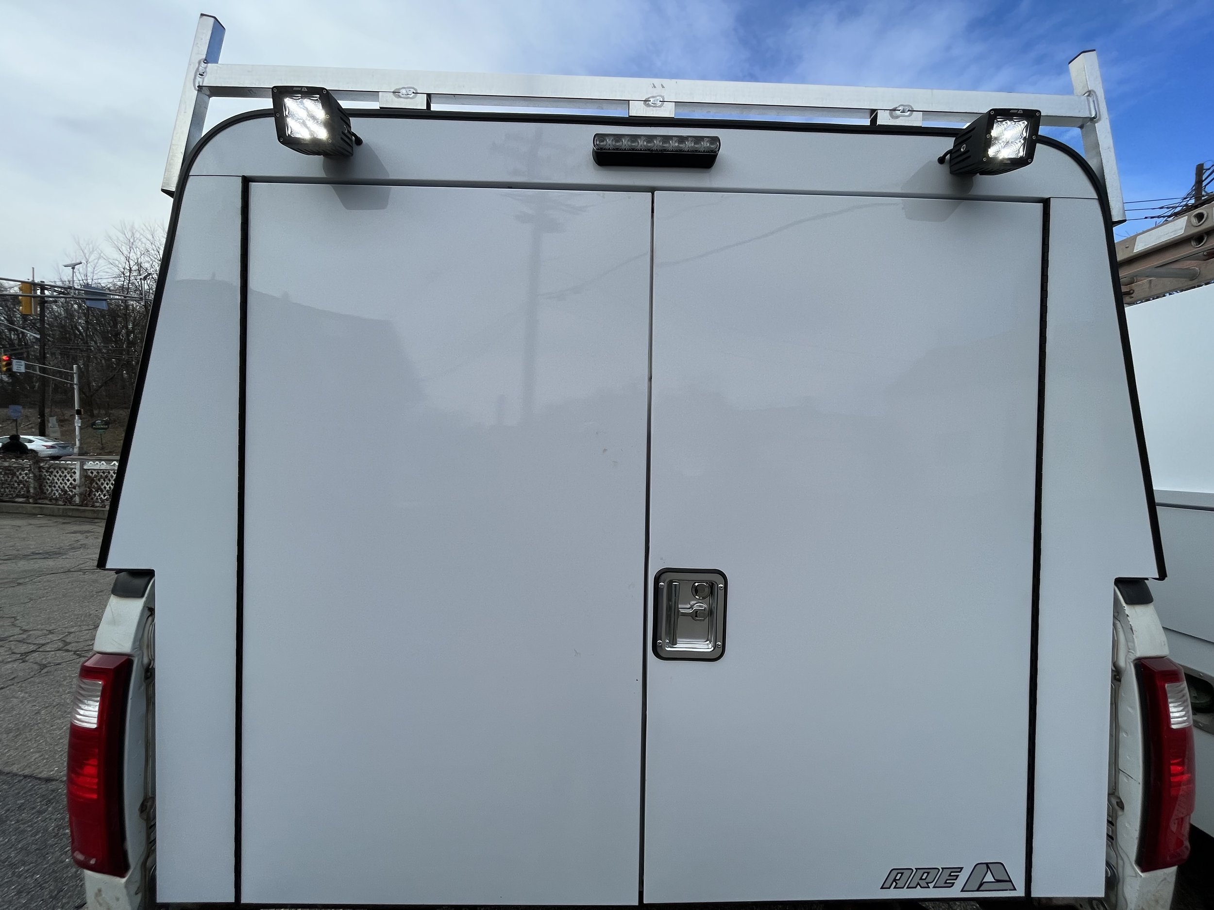 The rear view of a white utility or service truck with double doors, a handle on the right door, dual tail lights, and mounted work lights on the top corners. Full Size commercial DCU cap and full door that replaces truck tailgate.
