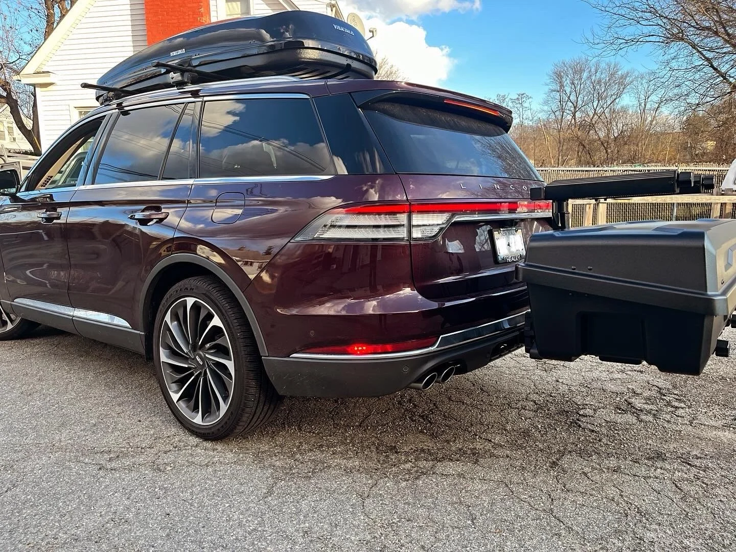 A maroon Lincoln SUV parked outdoors with a roof cargo box, a trailer hitch carrier, and dual exhaust pipes. House and trees in the background, clear sky with some clouds. Yakima EXO Gearlocker. Gear Locker on SUV