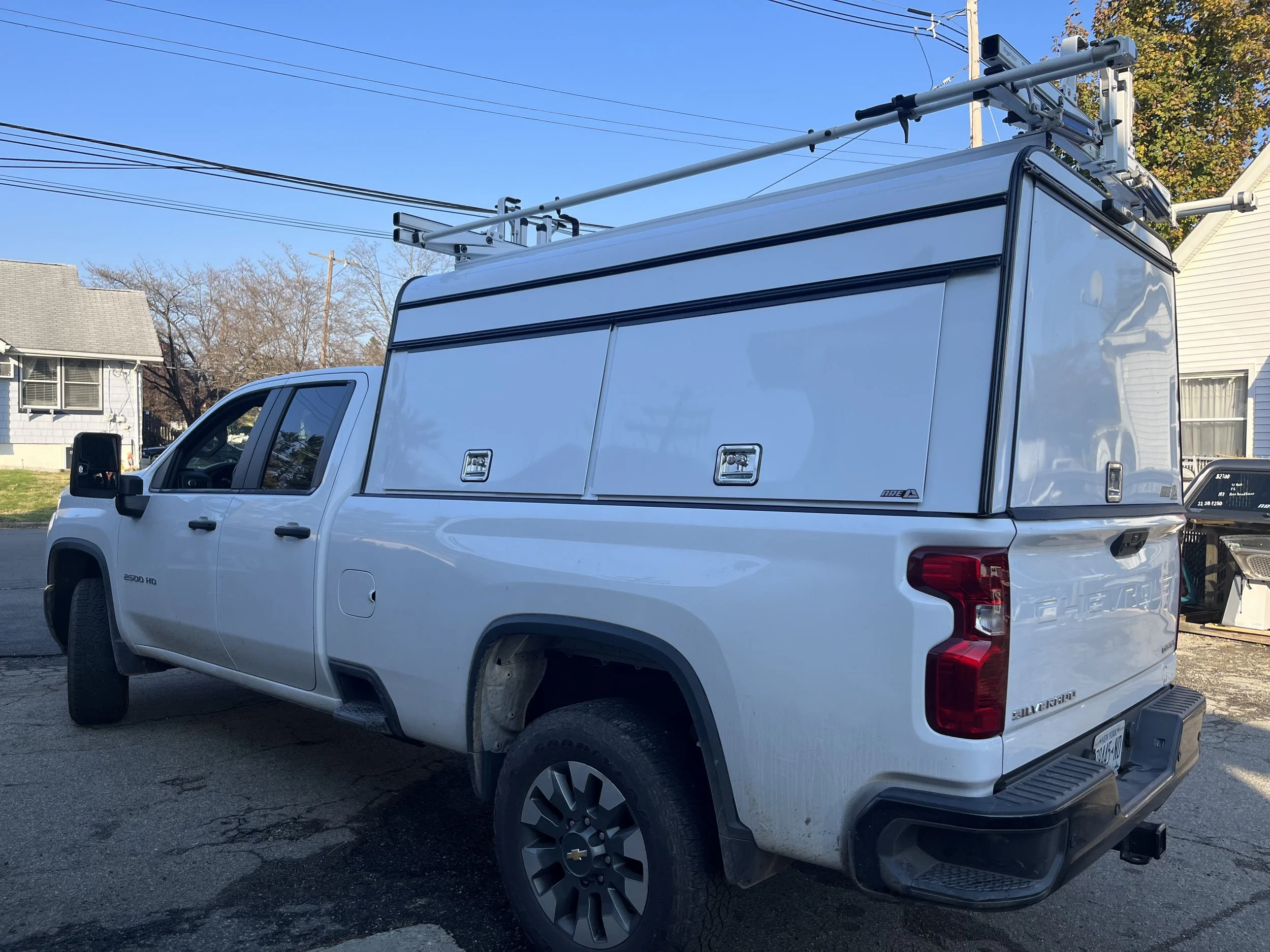White Chevrolet Silverado pickup truck with a custom camper shell and ladder rack in a residential area. 29" A.R.E Commercial DCU cap on a white Chevy.