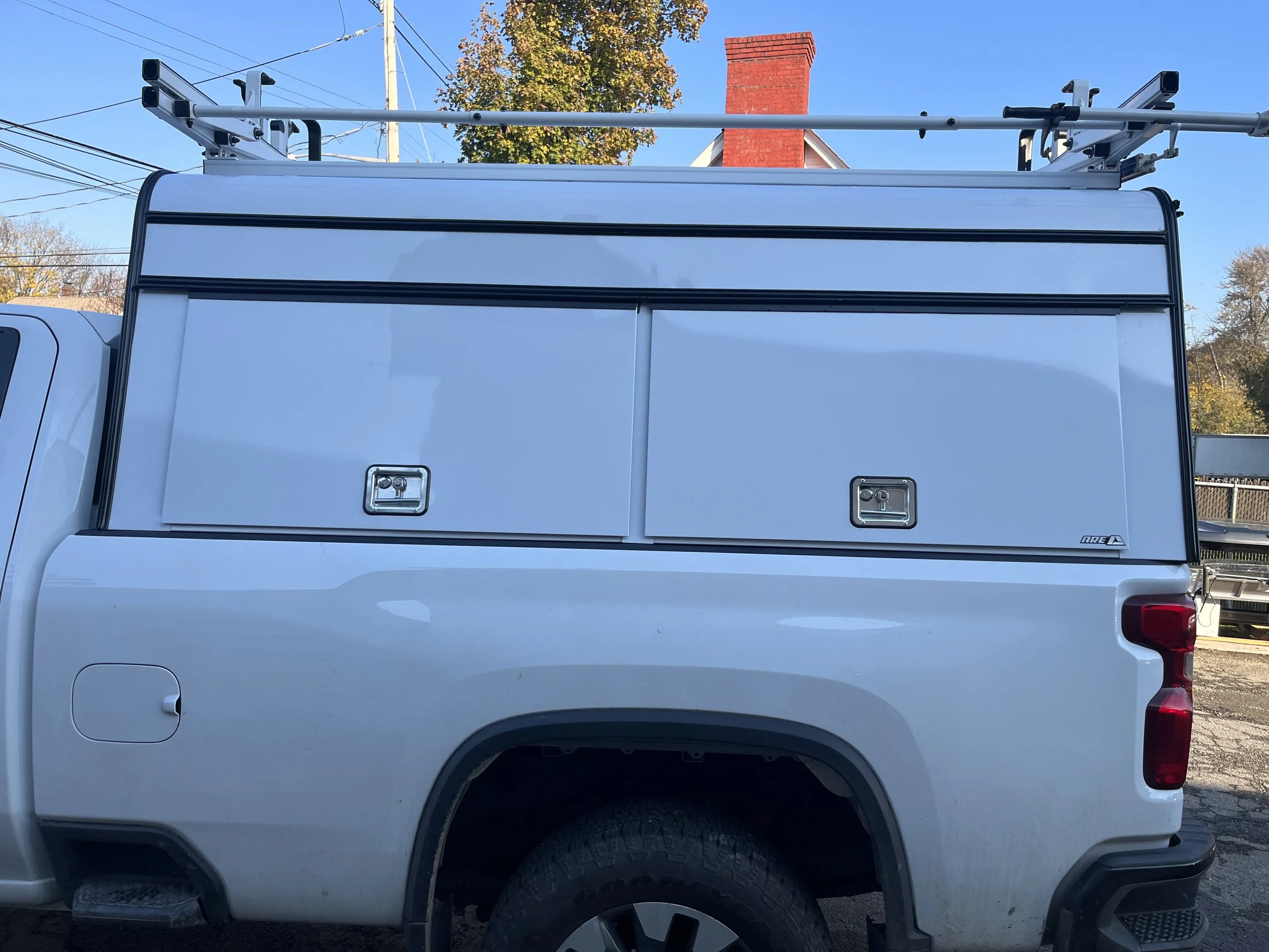 Side view of a white utility truck with a large enclosed storage compartment on the back, featuring two metal doors with handles and locks. A ladder rack is mounted on the top of the compartment.  29" A.R.E Commercial DCU cap on a white Chevy.