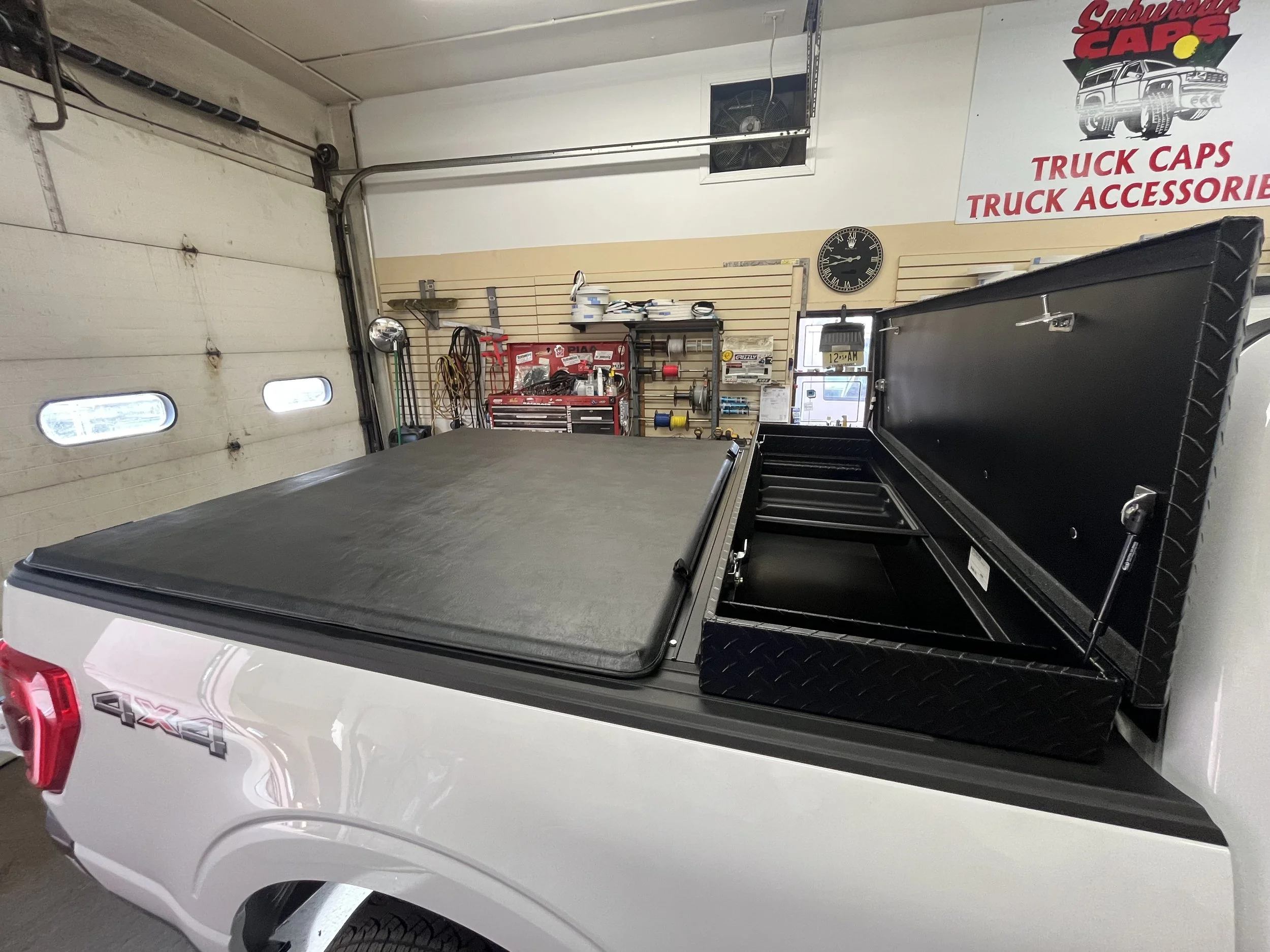 A white pickup truck with a black truck cap in a garage. The garage has tools, a wall clock, and signage for truck accessories on the wall. Ford F-150 with a tonneau cover and toolbox combo. Custom tonneau cover measure to size.