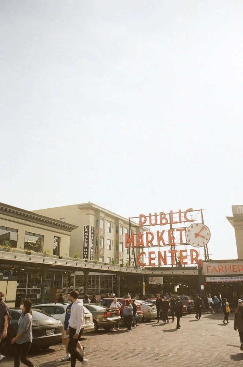 People walking in front of a public market center with a large sign and clock on the building.