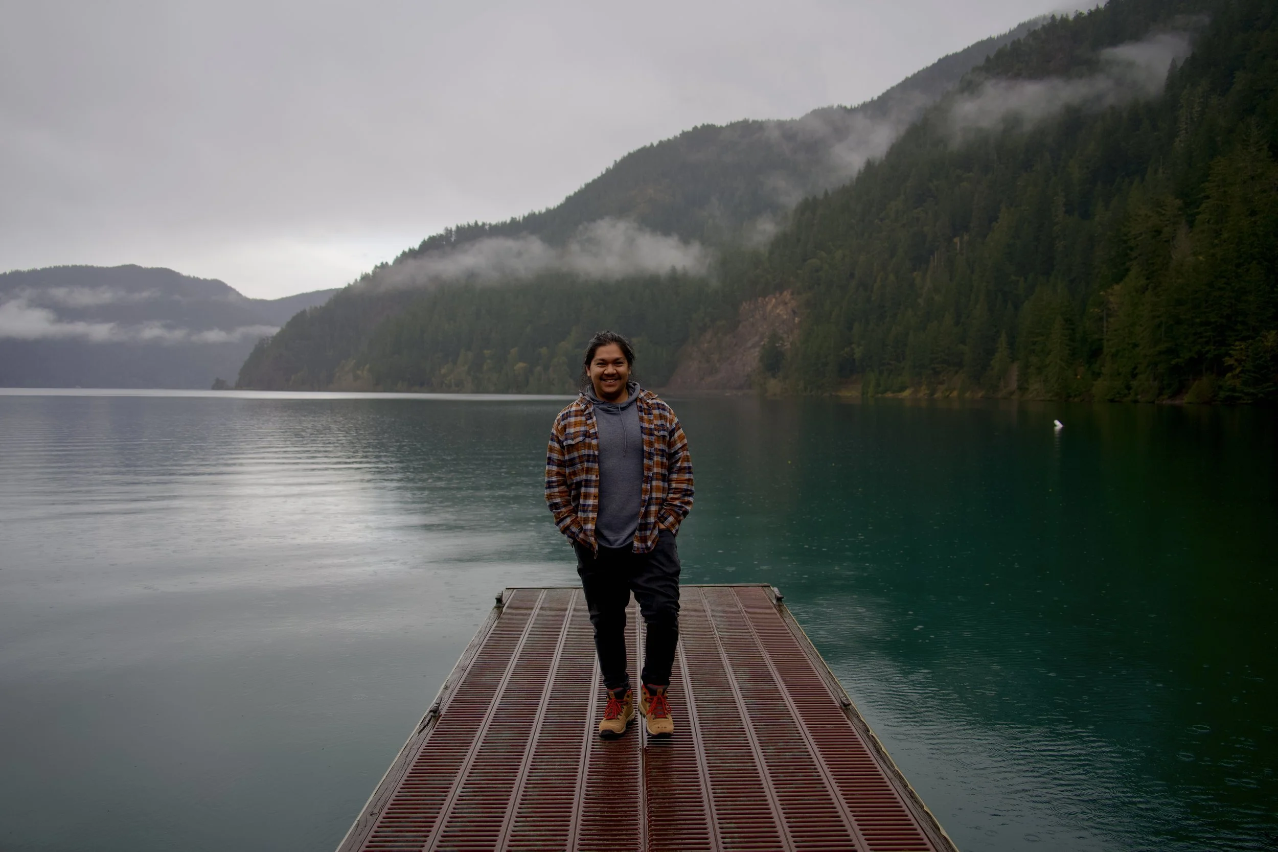 A person smiling and standing on a dock by a calm lake with forested hills and low clouds in the background on a cloudy day.