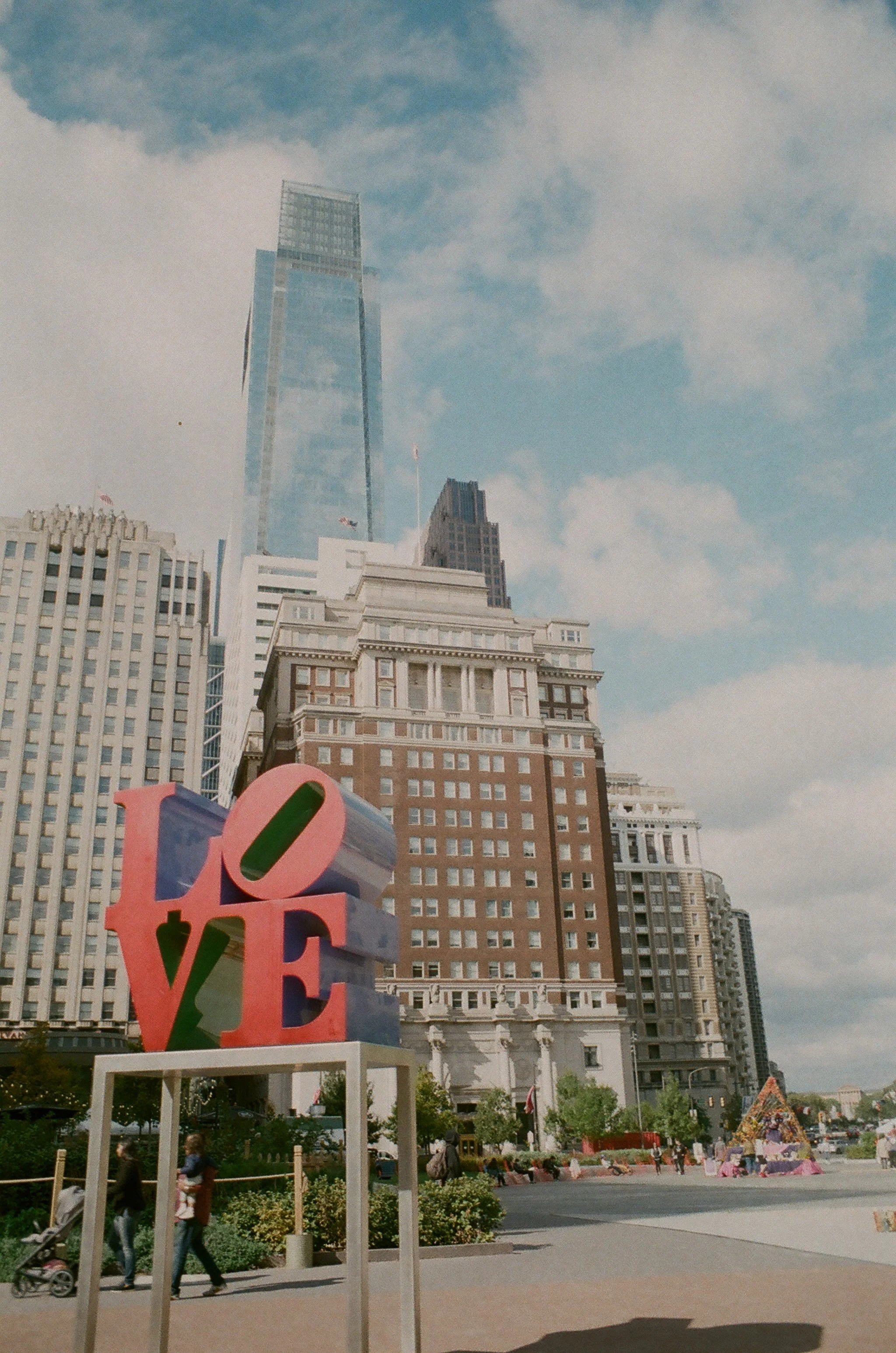 Cityscape with tall buildings, a large colorful LOVE sculpture in the foreground, and a blue sky with clouds.