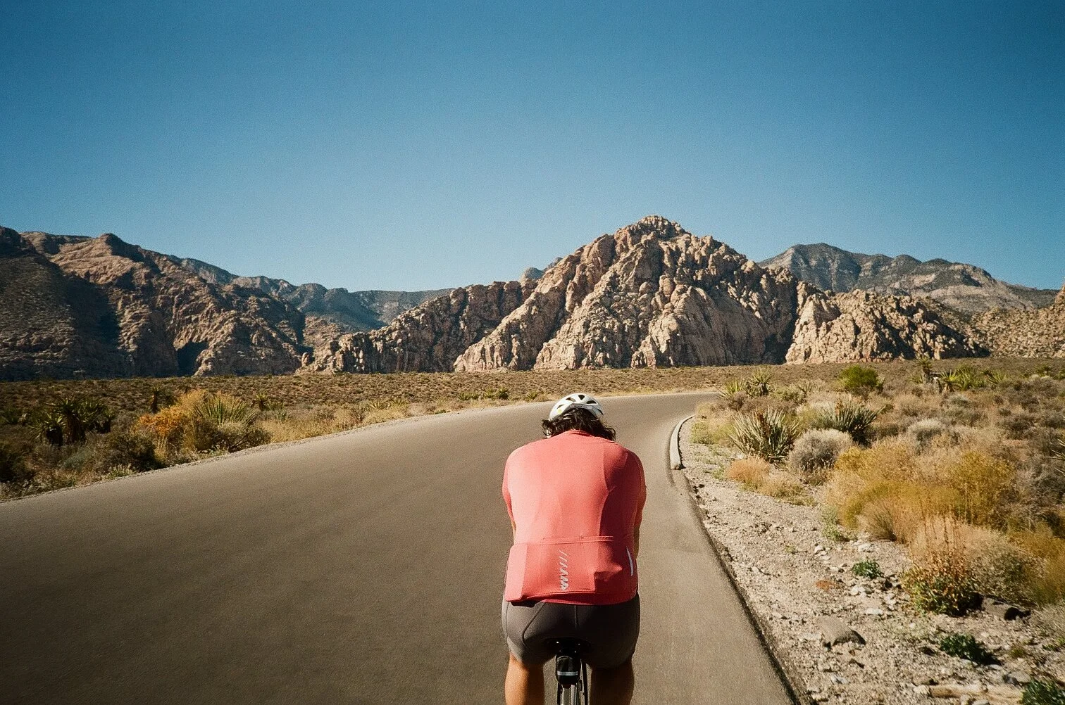 A person in a pink shirt and black shorts riding a bicycle on a desert road with mountains in the background.