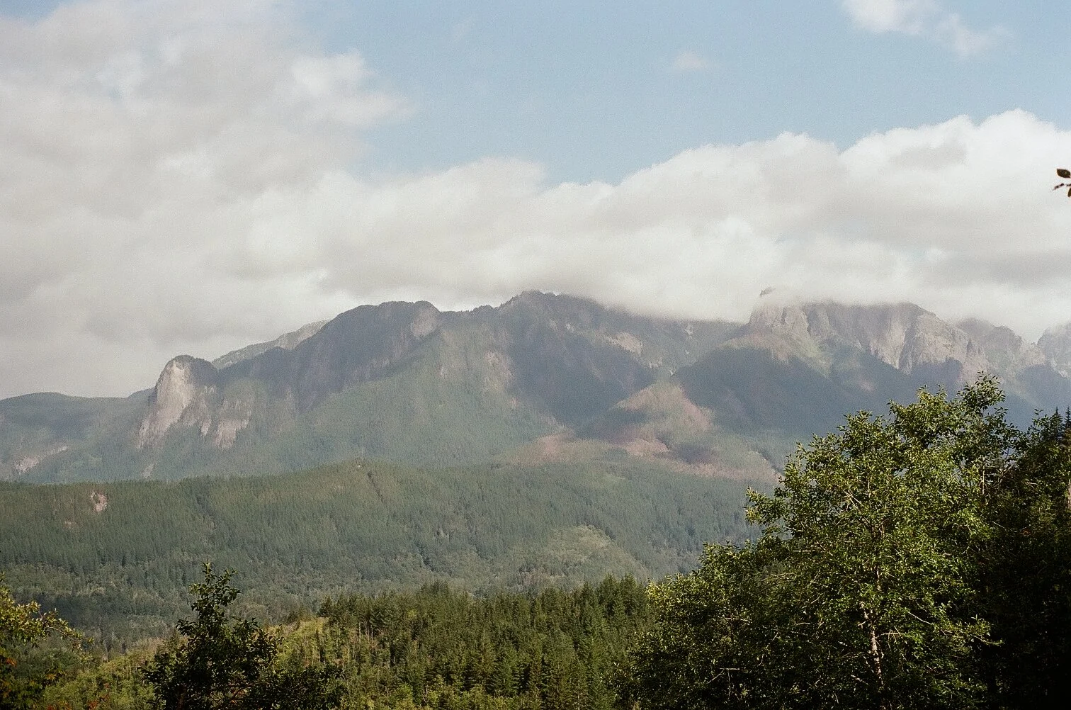 A mountain range with lush green forested slopes, partially covered by clouds under a partly cloudy sky.