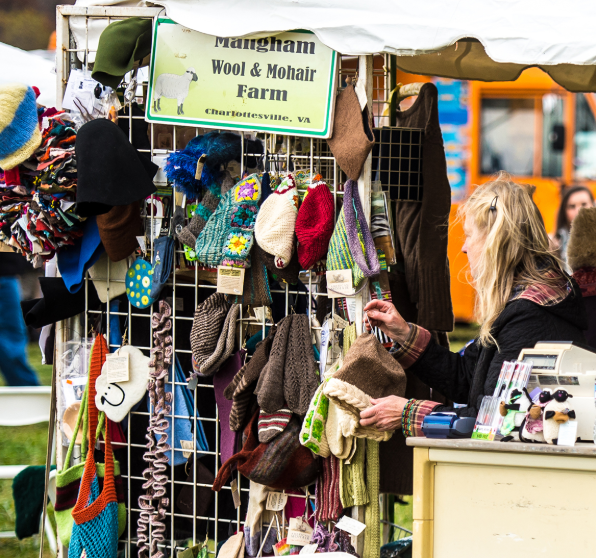A woman shopping at a market stall displaying wool and mohair hats, gloves, and scarves from Mangham Wool & Mohair Farm in Charlottesville, Virginia.