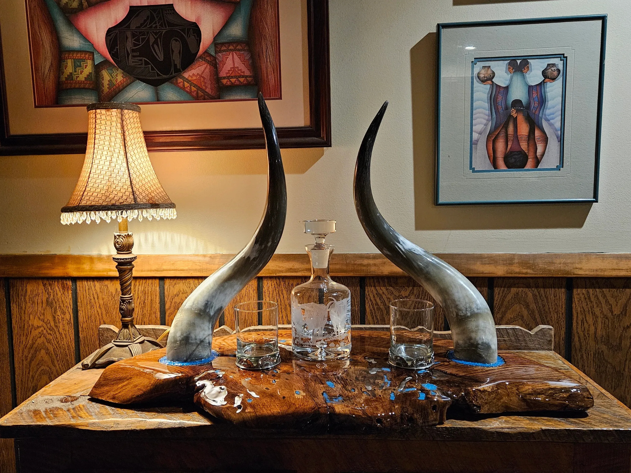 Decorative table with two curved horns, a glass decanter, and two glasses on a wooden surface. There are framed artwork on the wall and a lit table lamp in the background.