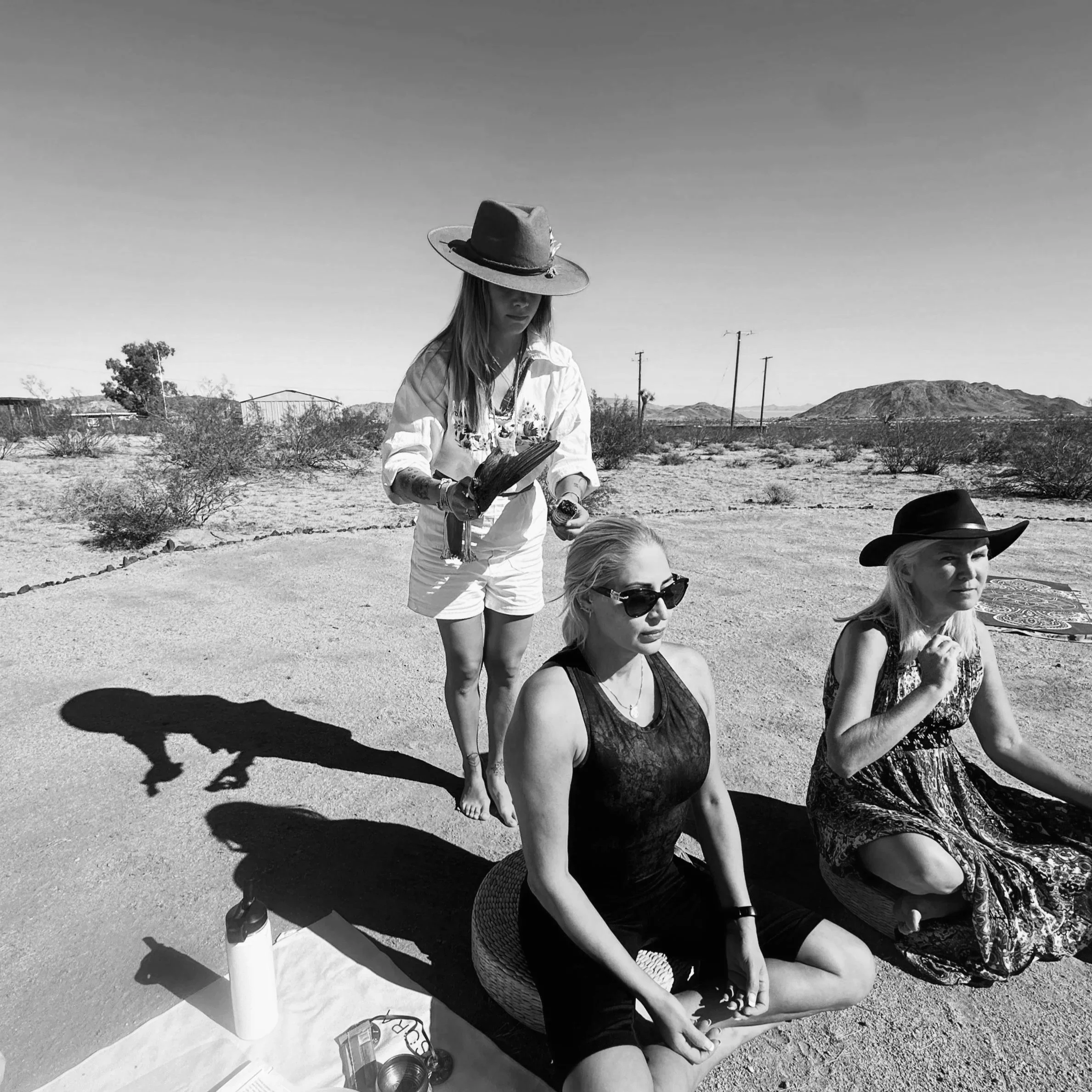 Three women practicing yoga outdoors in a desert landscape, two sitting cross-legged and one standing. All wear hats, with one woman holding a notebook, and the scene is captured in black and white.