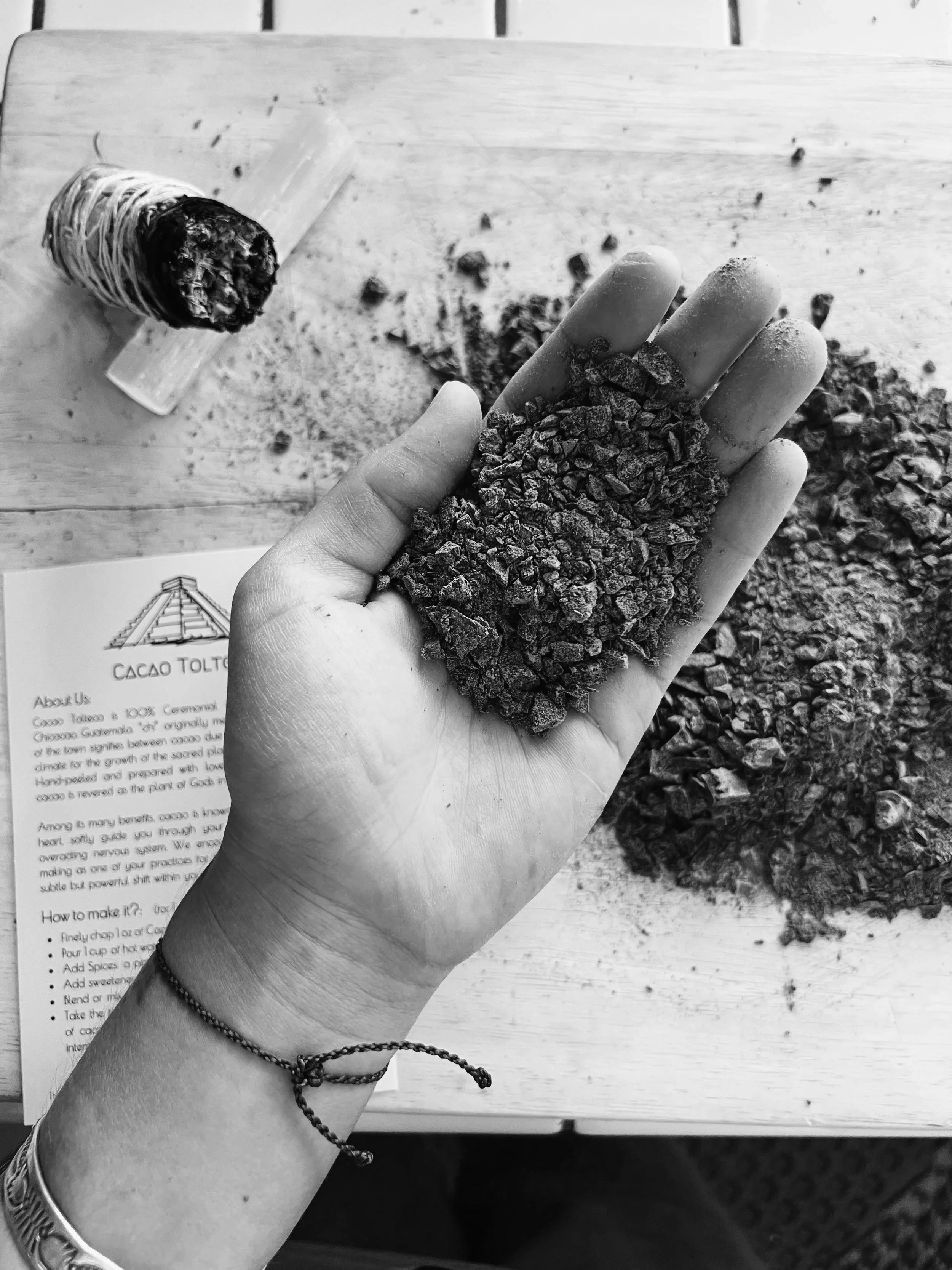A hand holding crushed cacao nibs over a wooden table with scattered nibs, a piece of paper with some text, and a cacao pod in the background.