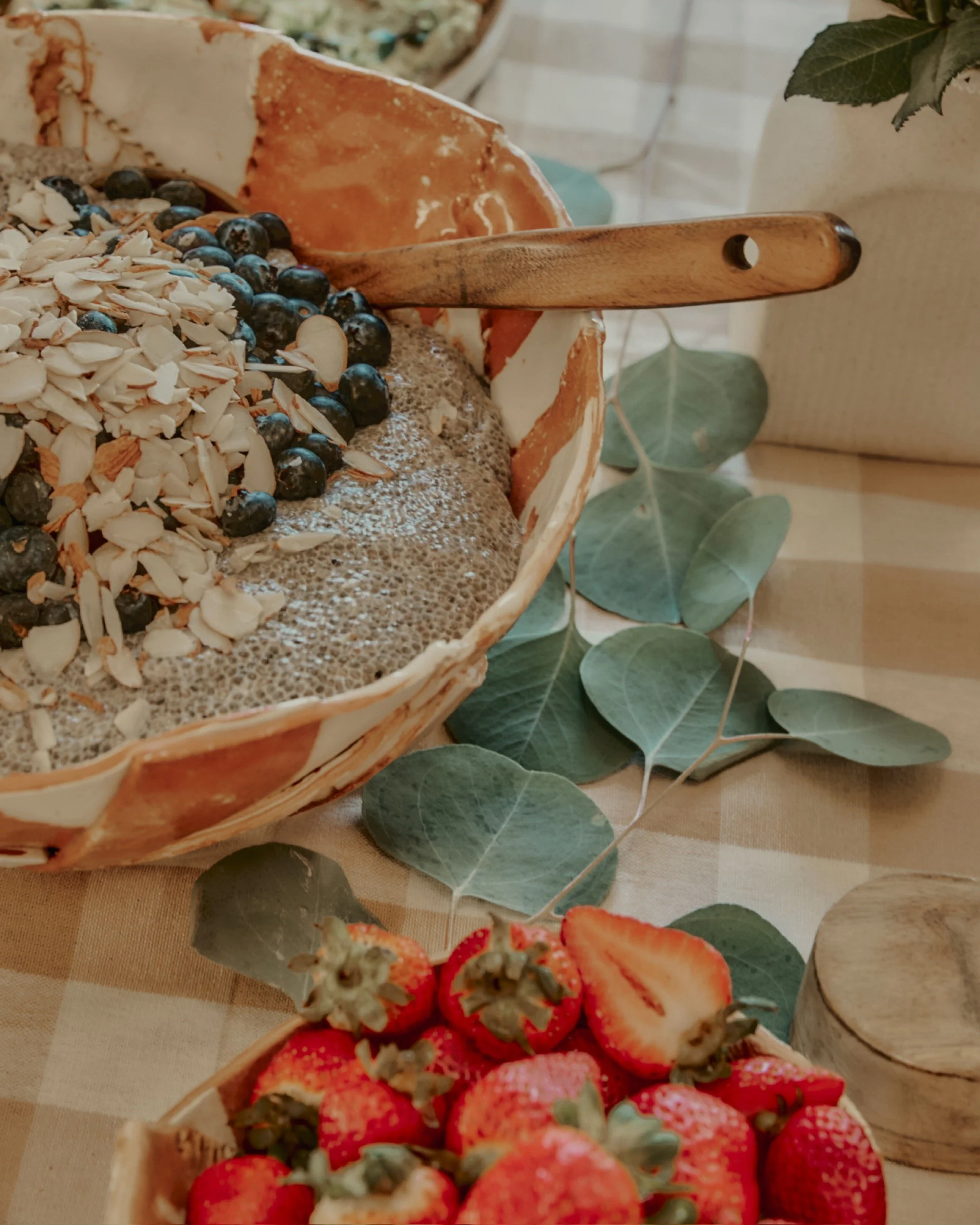 A rustic picnic table with a checkered tablecloth, featuring a woven basket of strawberries, a pie topped with blueberries and sliced almonds, and a wooden spreader with eucalyptus leaves as decoration.