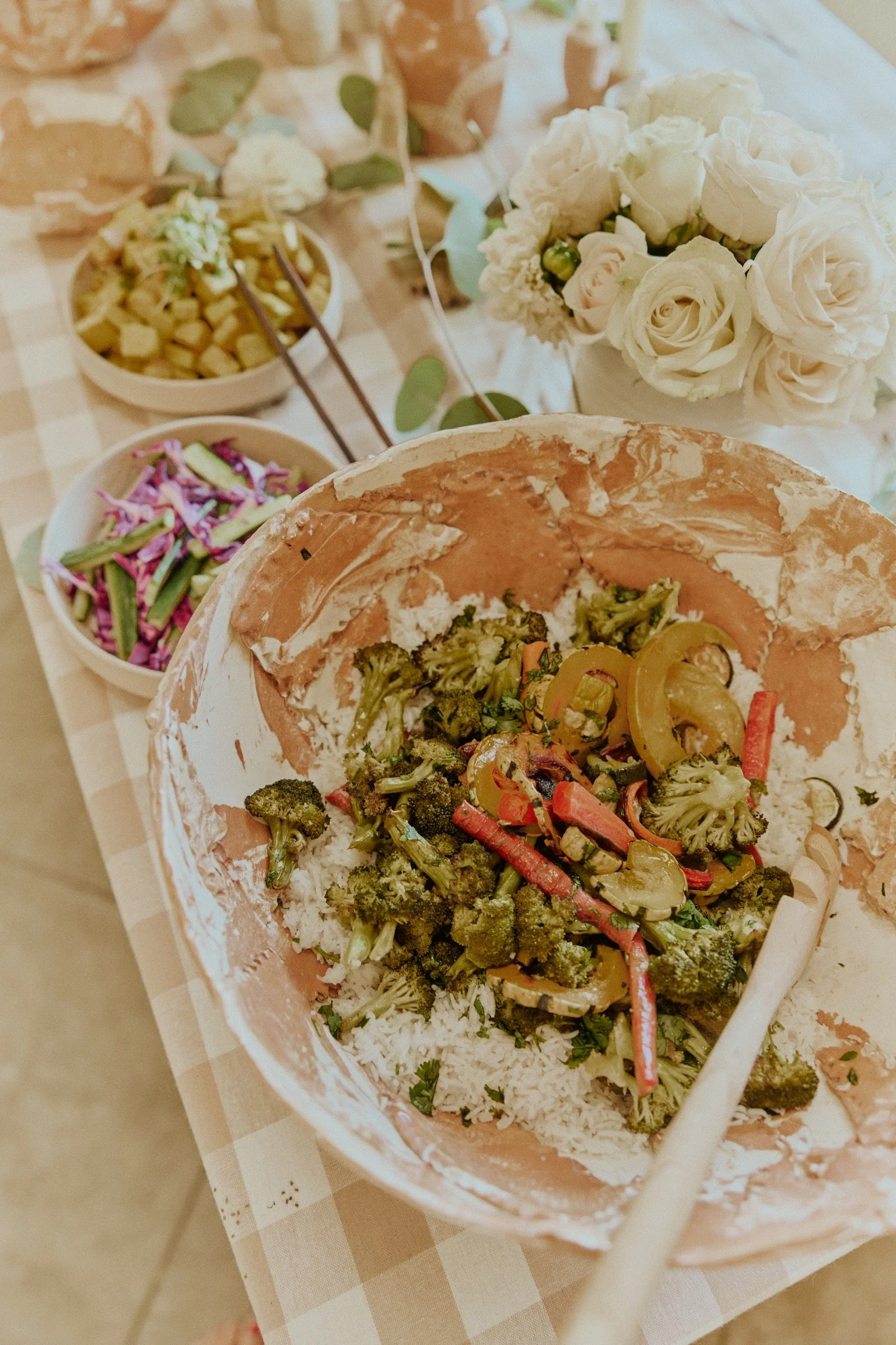 Empty salad bowl with broccoli and bell peppers, surrounded by bowls of purple cabbage and cucumber salad, on a checkered tablecloth with white flower arrangement.