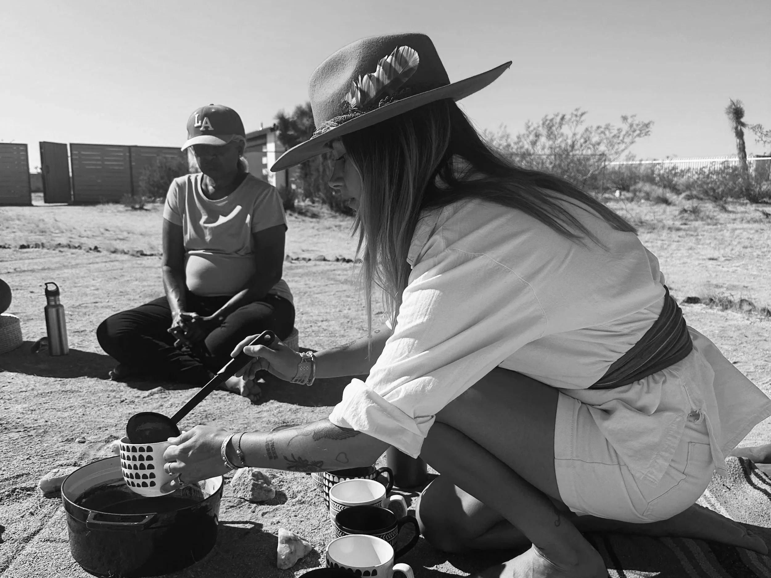 A woman with a wide-brimmed hat and tattoos, preparing coffee outdoors while sitting on a rug in a desert landscape. A woman in a LA baseball cap is sitting nearby.