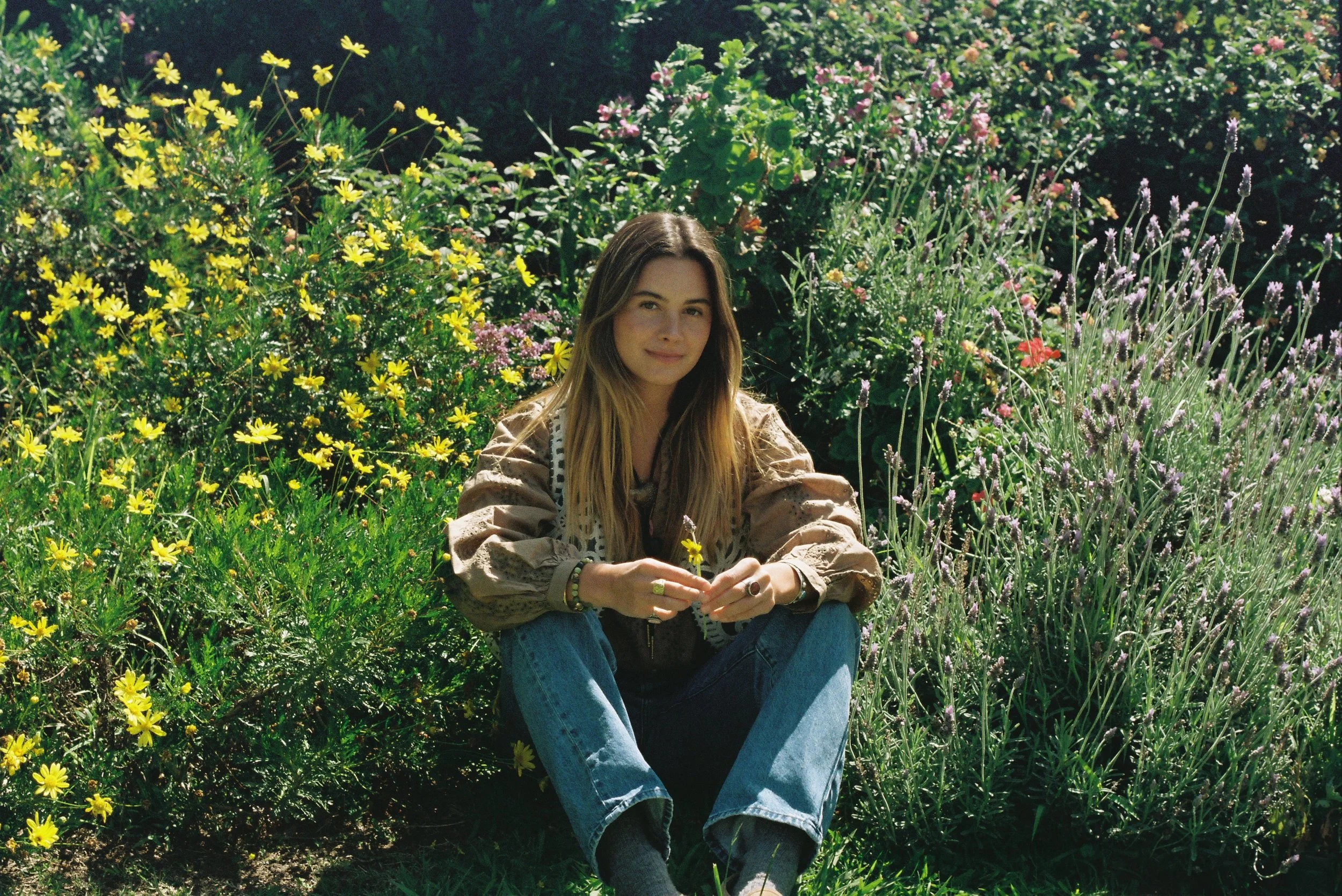 A young woman with long hair sitting in a garden surrounded by yellow and purple flowers.
