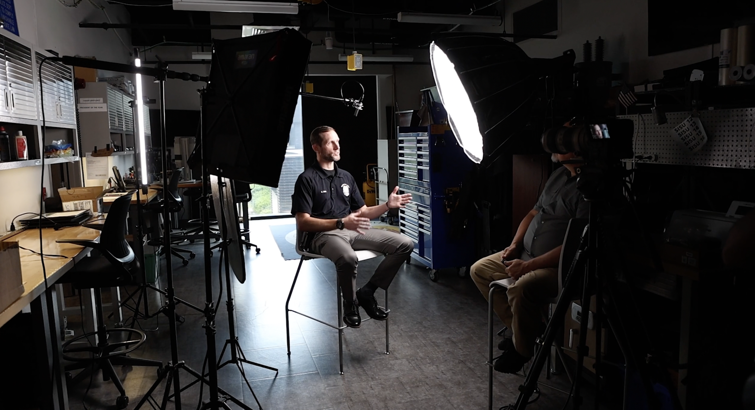 A man in a black shirt and khakis is sitting on a chair, gesturing with his hands, being recorded for an interview or presentation in a studio with lighting equipment, camera, and a person operating the camera.