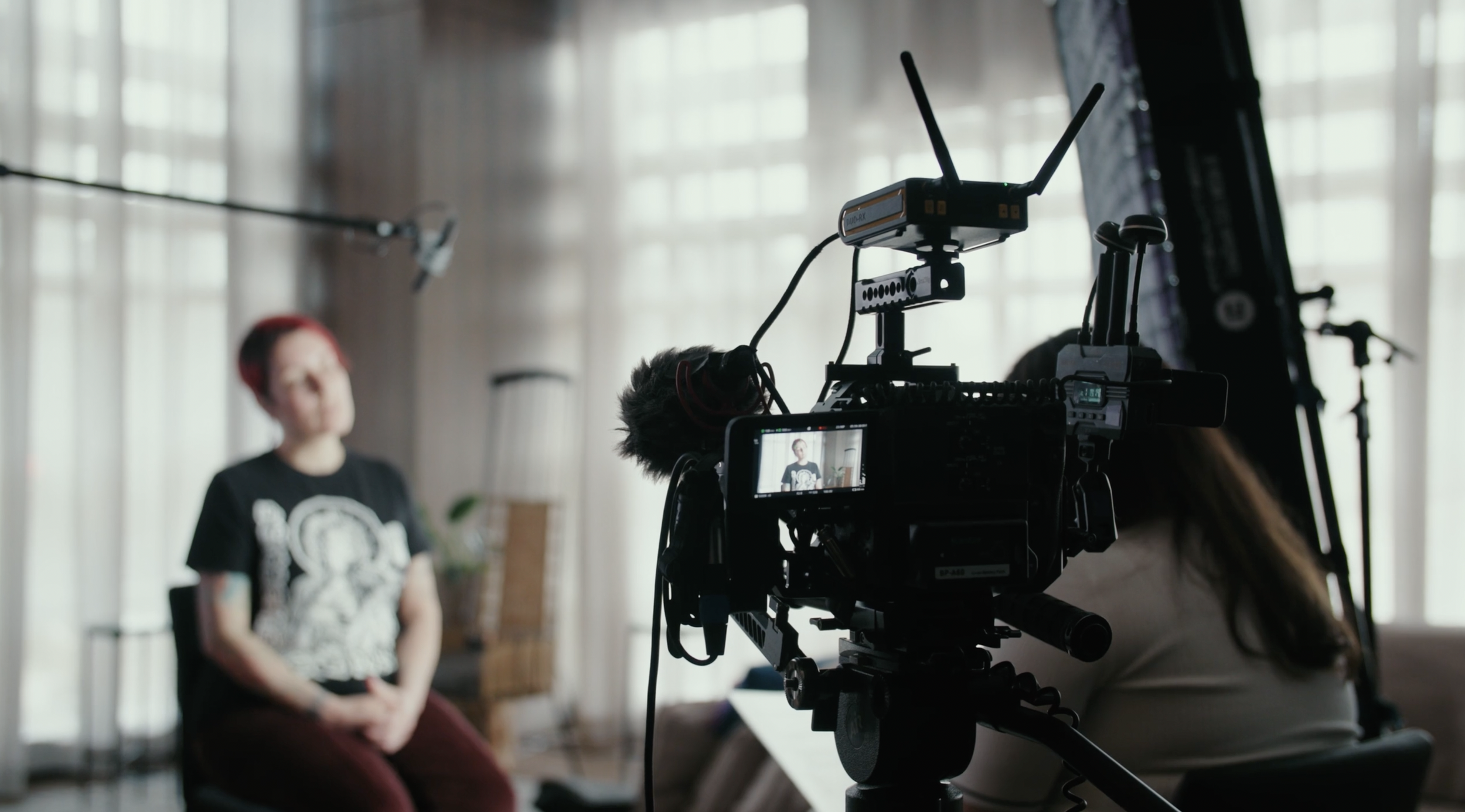 A video camera records a woman with red hair wearing a black T-shirt, sitting on a chair with her hands in her lap, in a softly lit room with large windows and sheer curtains, while a boom microphone hovers above.