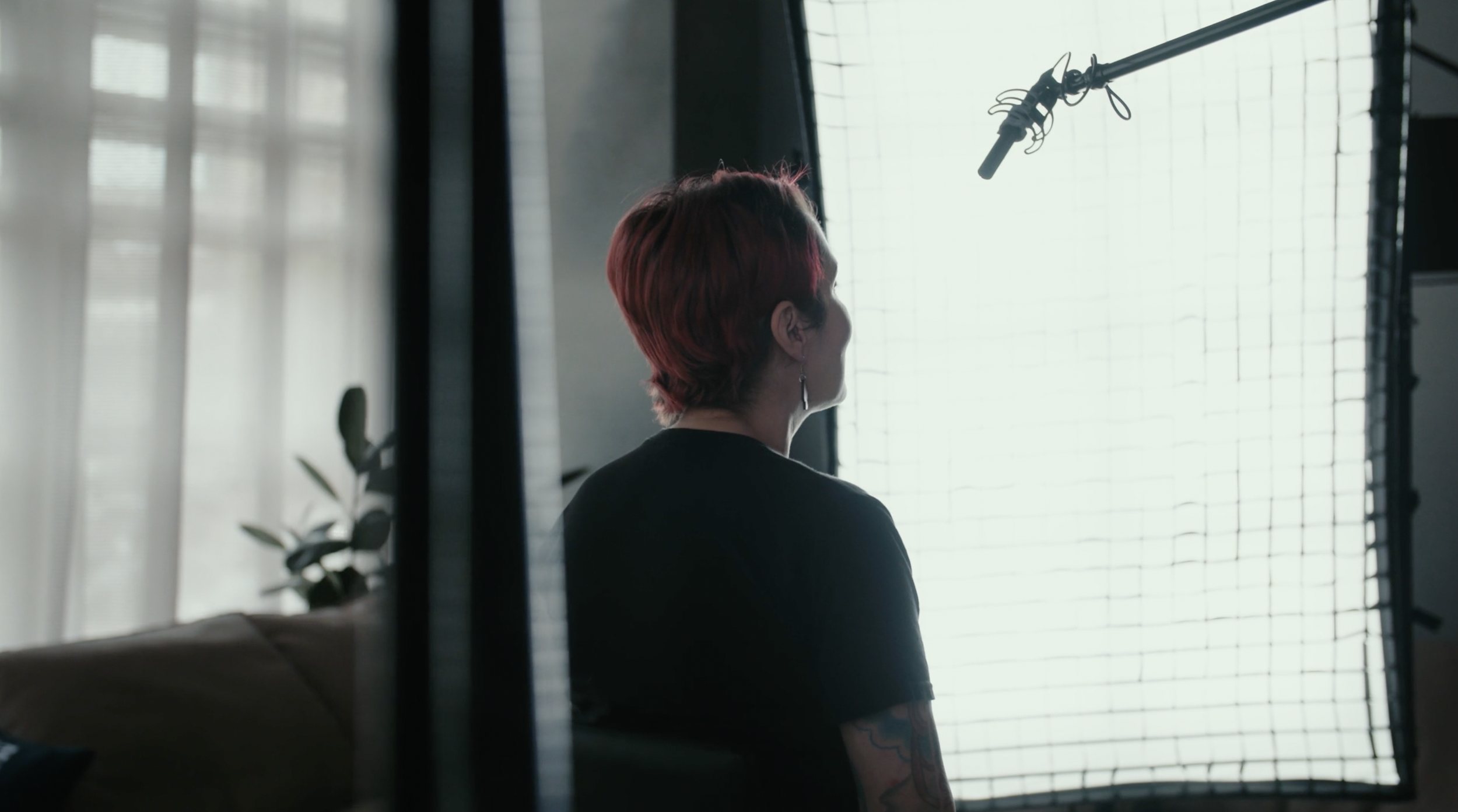 A person with short, reddish hair and tattoos on their arm, sitting in front of a large window with a grid pattern, during a filming or recording session with a microphone overhead.
