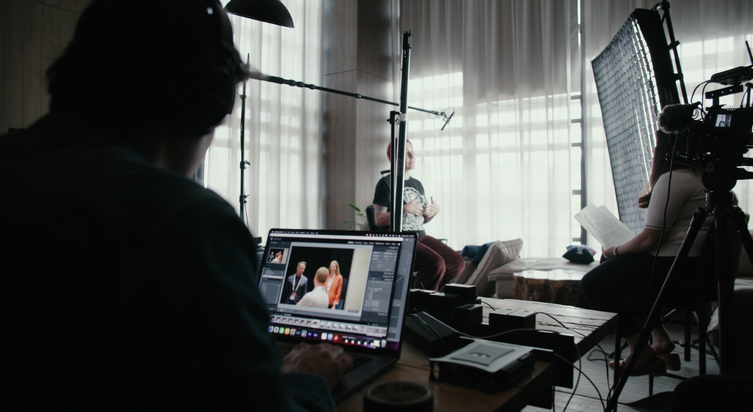 A behind-the-scenes view of a video or photo shoot, showing a woman speaking in front of a camera setup, with crew members operating equipment, including a laptop and lighting, in a room with large windows and sheer curtains.