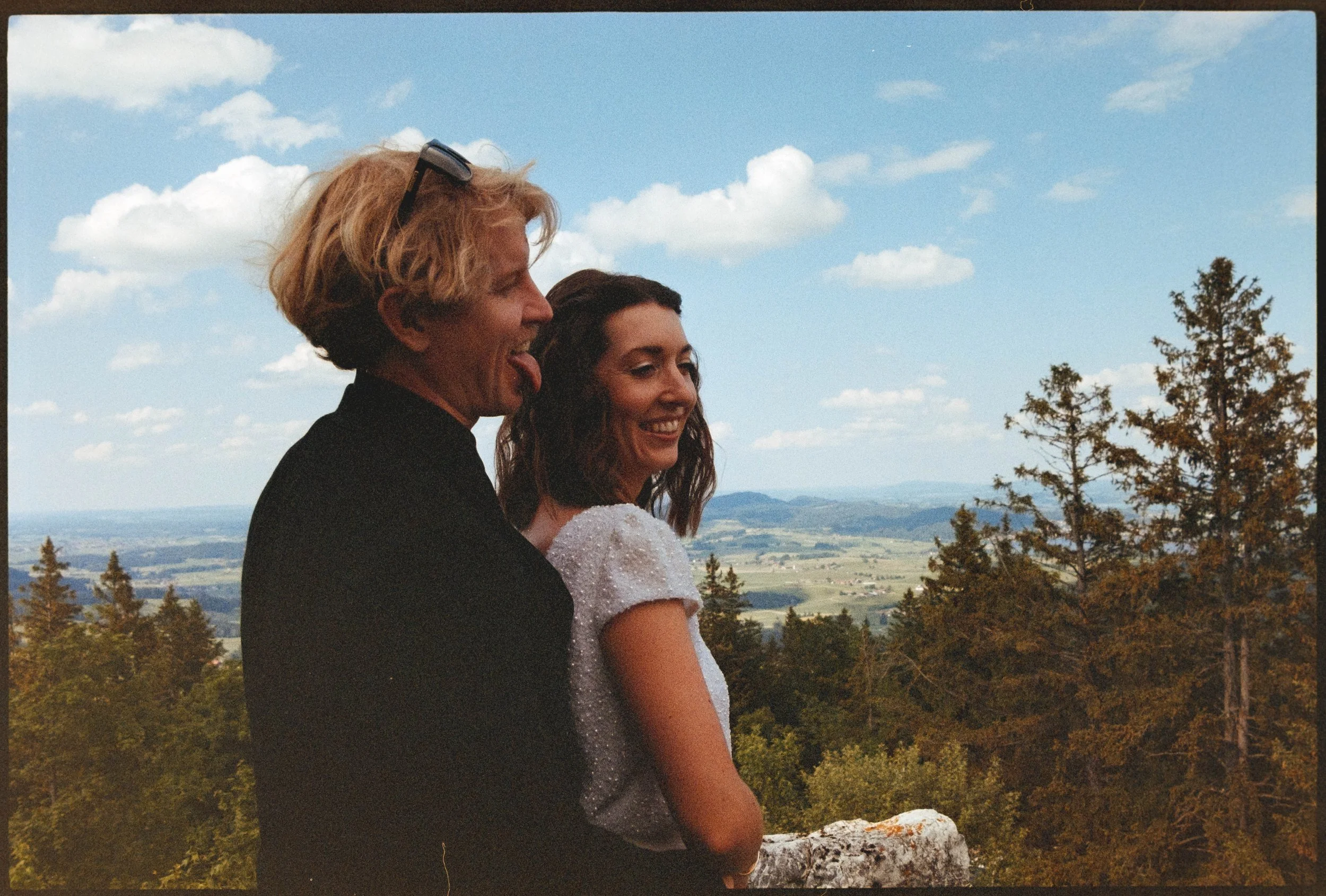 A man and a woman sitting outdoors on a rock, with a scenic background of trees, hills, and a blue sky with clouds.