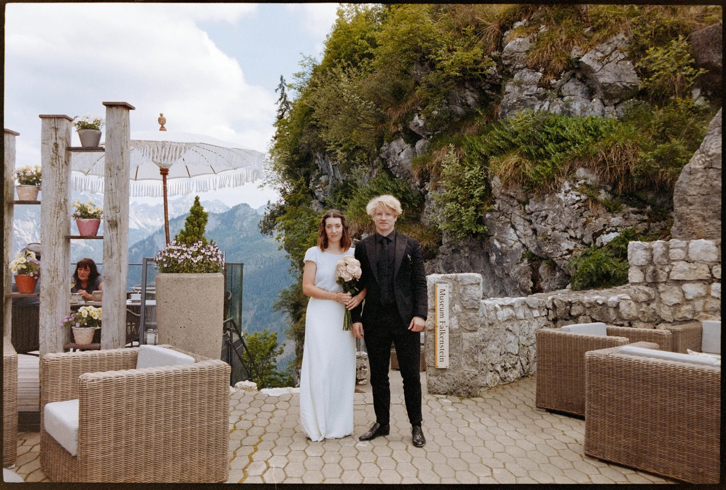 Eine Braut und ein Bräutigam stehen auf einer Terrasse mit Bergen im Hintergrund. Die Braut trägt ein weißes Kleid und hält einen Strauß rosa Rosen. Der Bräutigam ist in einen schwarzen Anzug gekleidet. Ein Schild mit Aufschrift „Museum Falkenstein"