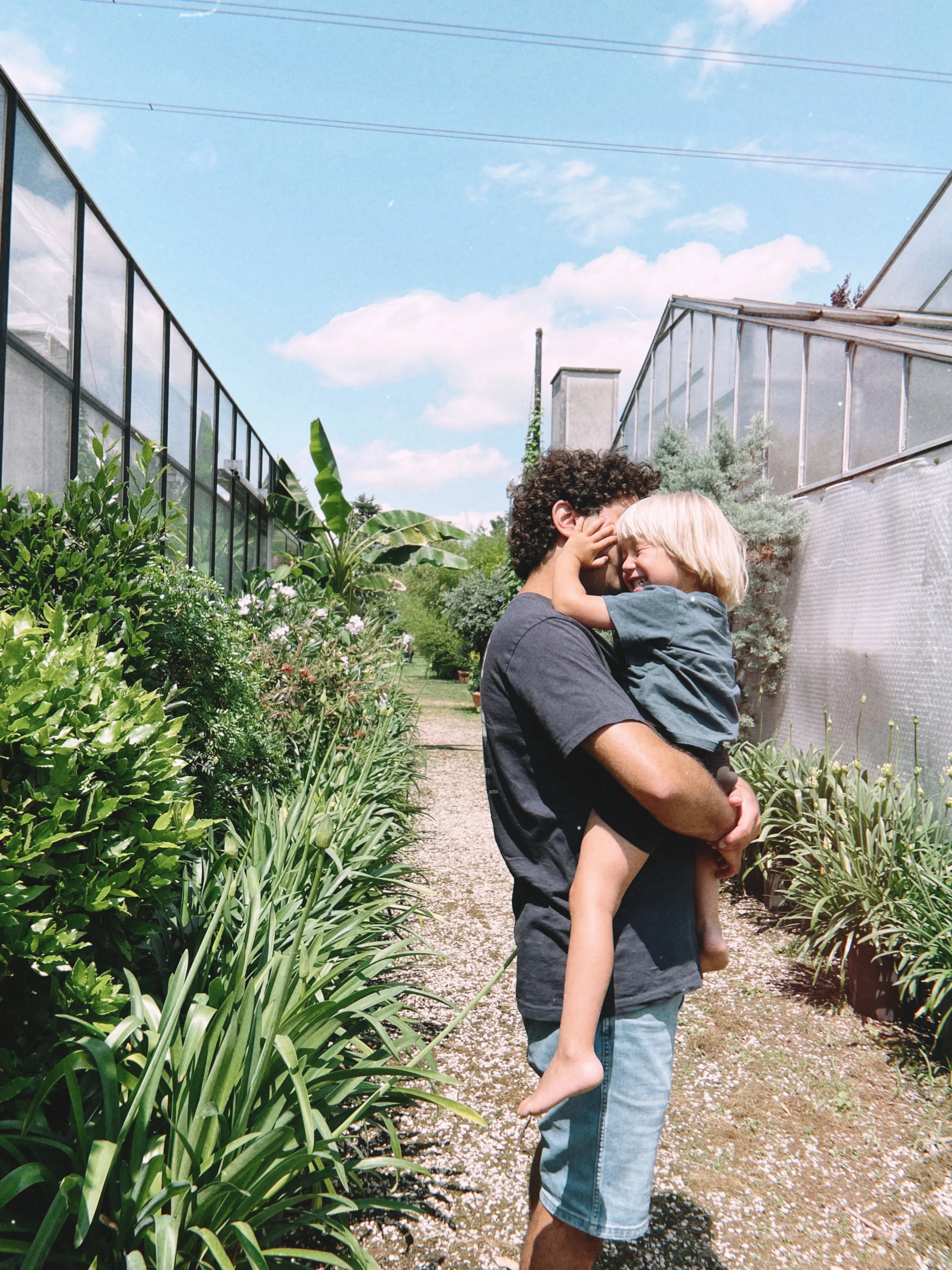 A man holding a young child in a garden with green plants and a greenhouse, under a blue sky with clouds.