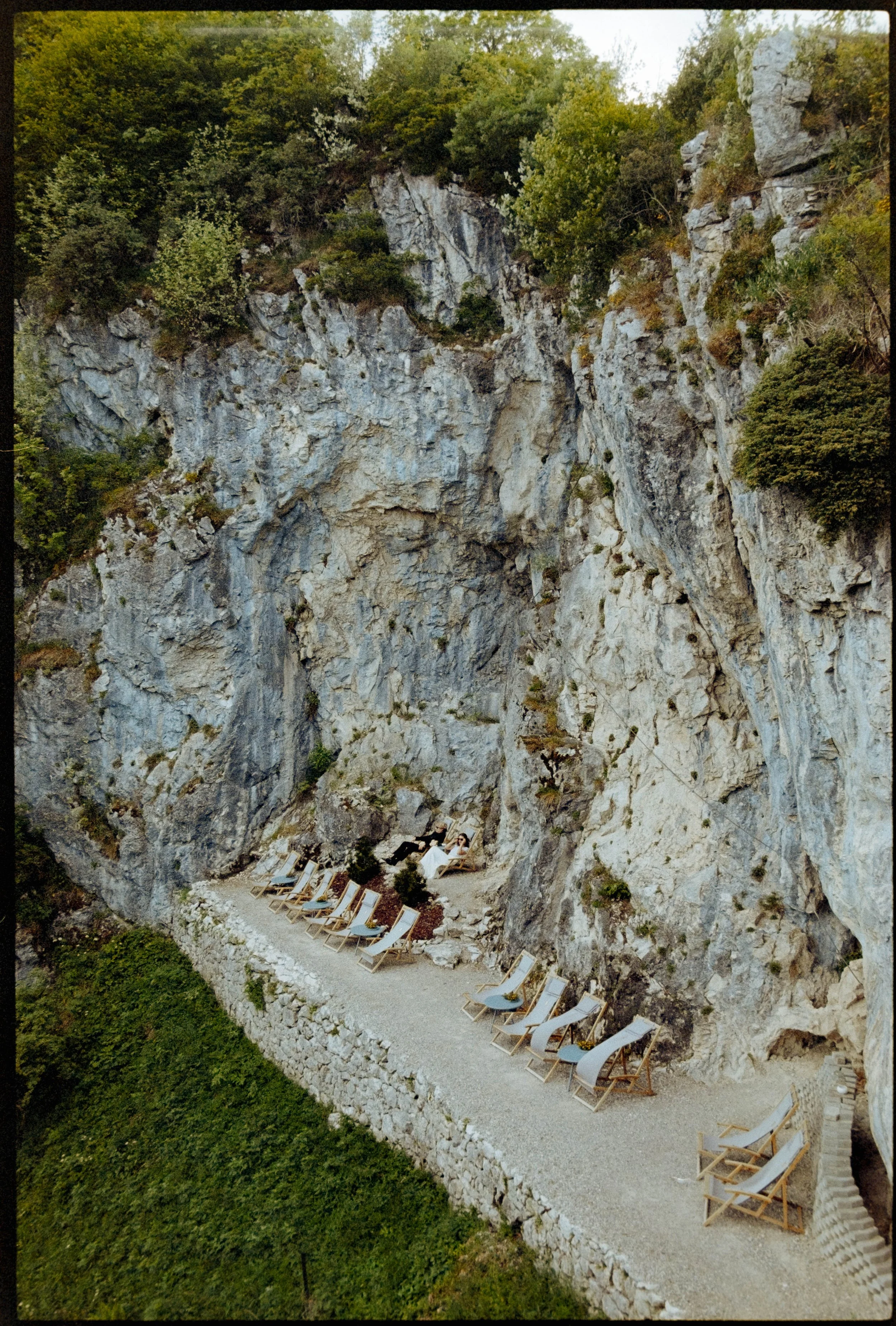 Couple lying on lounge chairs in a narrow gravel path beneath a rocky cliffside surrounded by green trees.