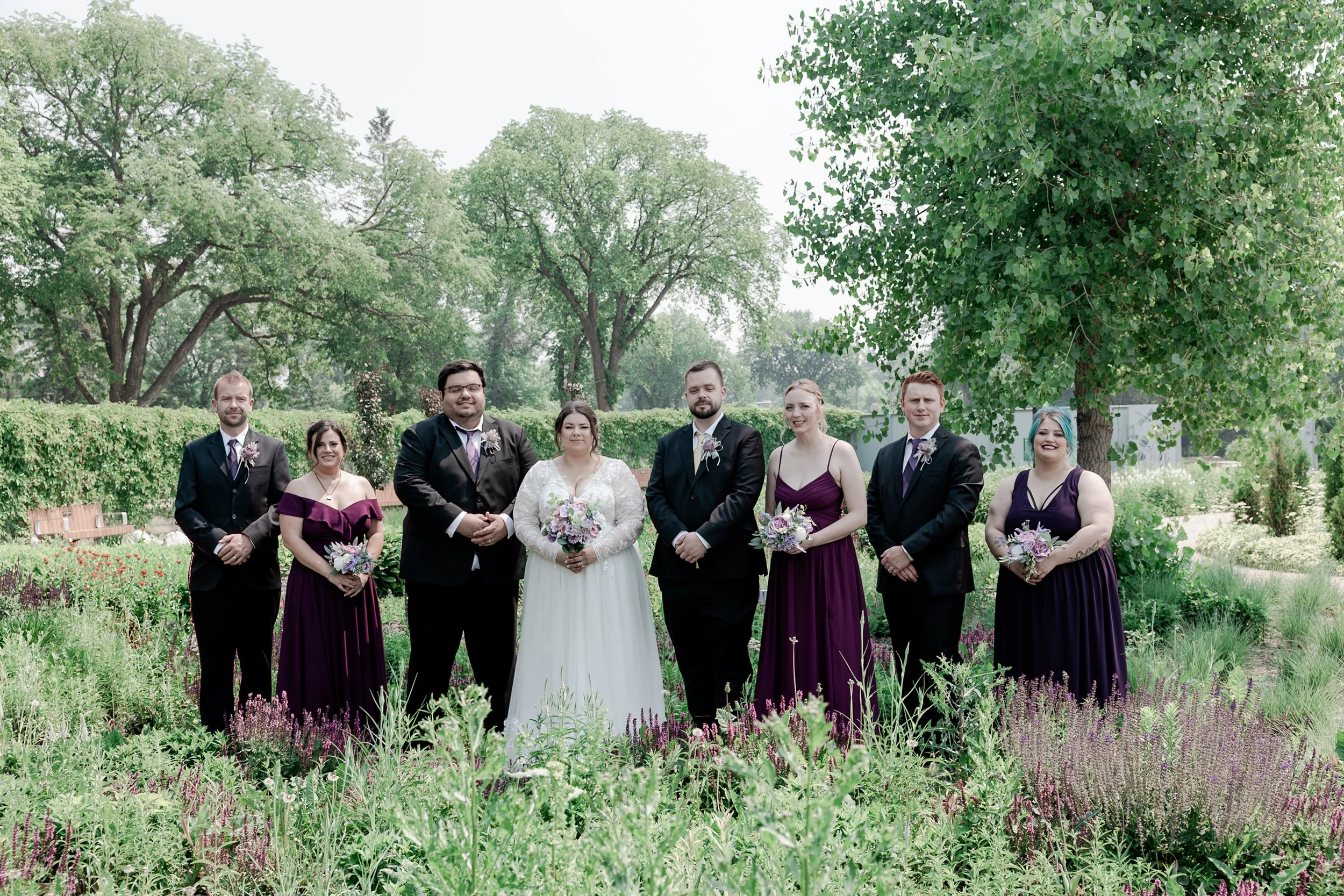 Wedding party standing in a lush garden with trees and flowering plants, including the bride in a white gown, bridesmaids in purple dresses, and groomsmen in black suits.