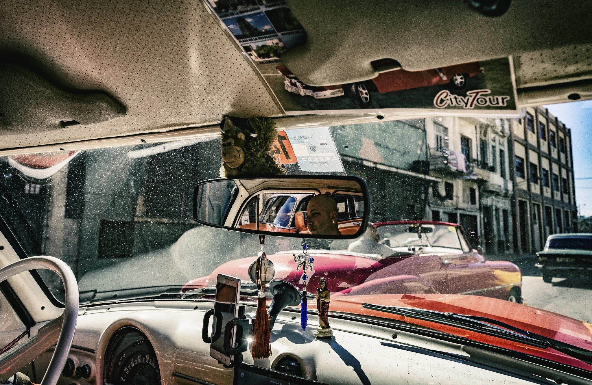 The mirror holds a face, the windshield holds the city, the dashboard holds the saints — and somehow it all moves forward together. © Bob Plotkin