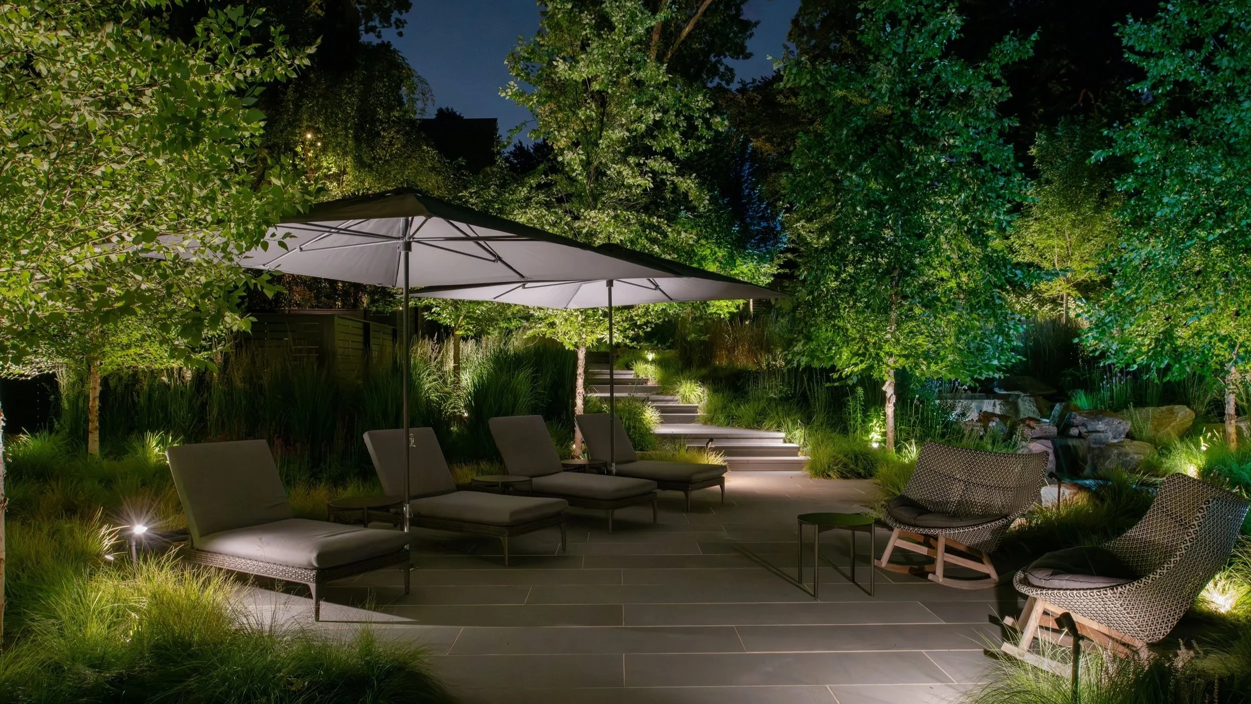 Nighttime terrace with lounge seating, umbrellas, soft LED path lighting, and illuminated trees creating depth and layered texture across a New Jersey garden.