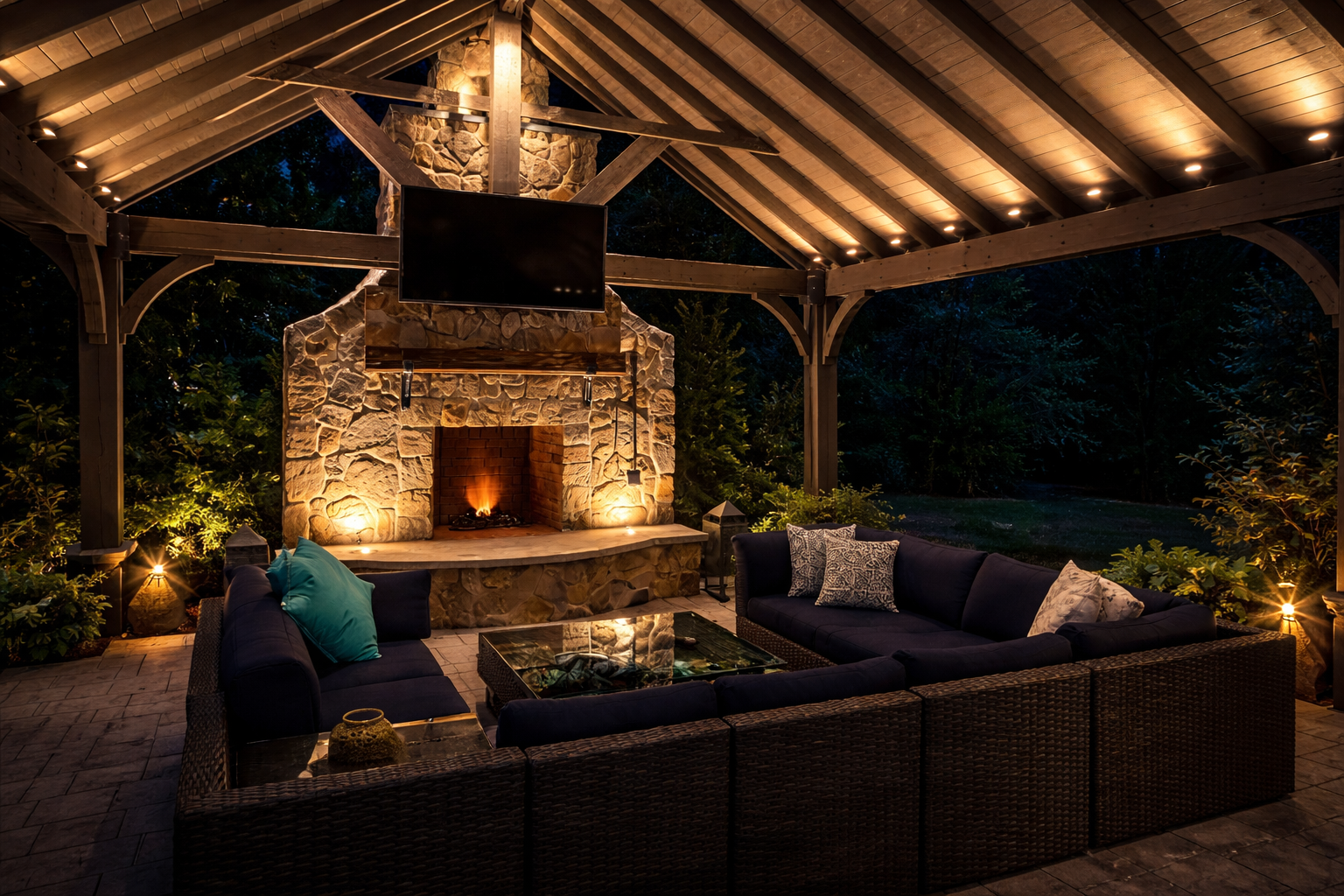 Covered outdoor pavilion in New Jersey featuring a stone fireplace and warm architectural landscape lighting, with soft downlighting in exposed wood beams and subtle uplighting that creates an inviting nighttime living space.